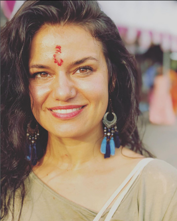 Close-up of a woman smiling with dark curly hair, wearing large earrings with blue tassels and a nose stud, outdoors with blurred colorful background.