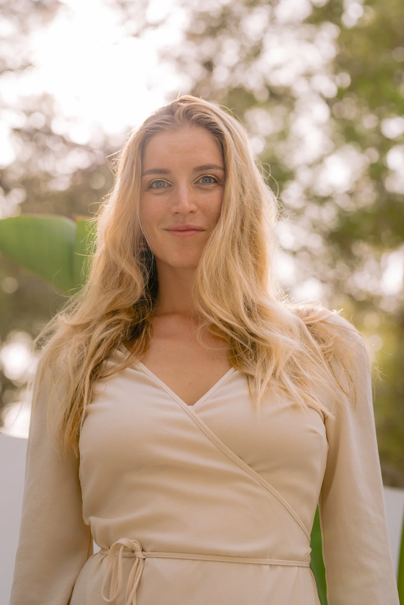 A smiling woman with long blonde hair standing outdoors with trees in the background.