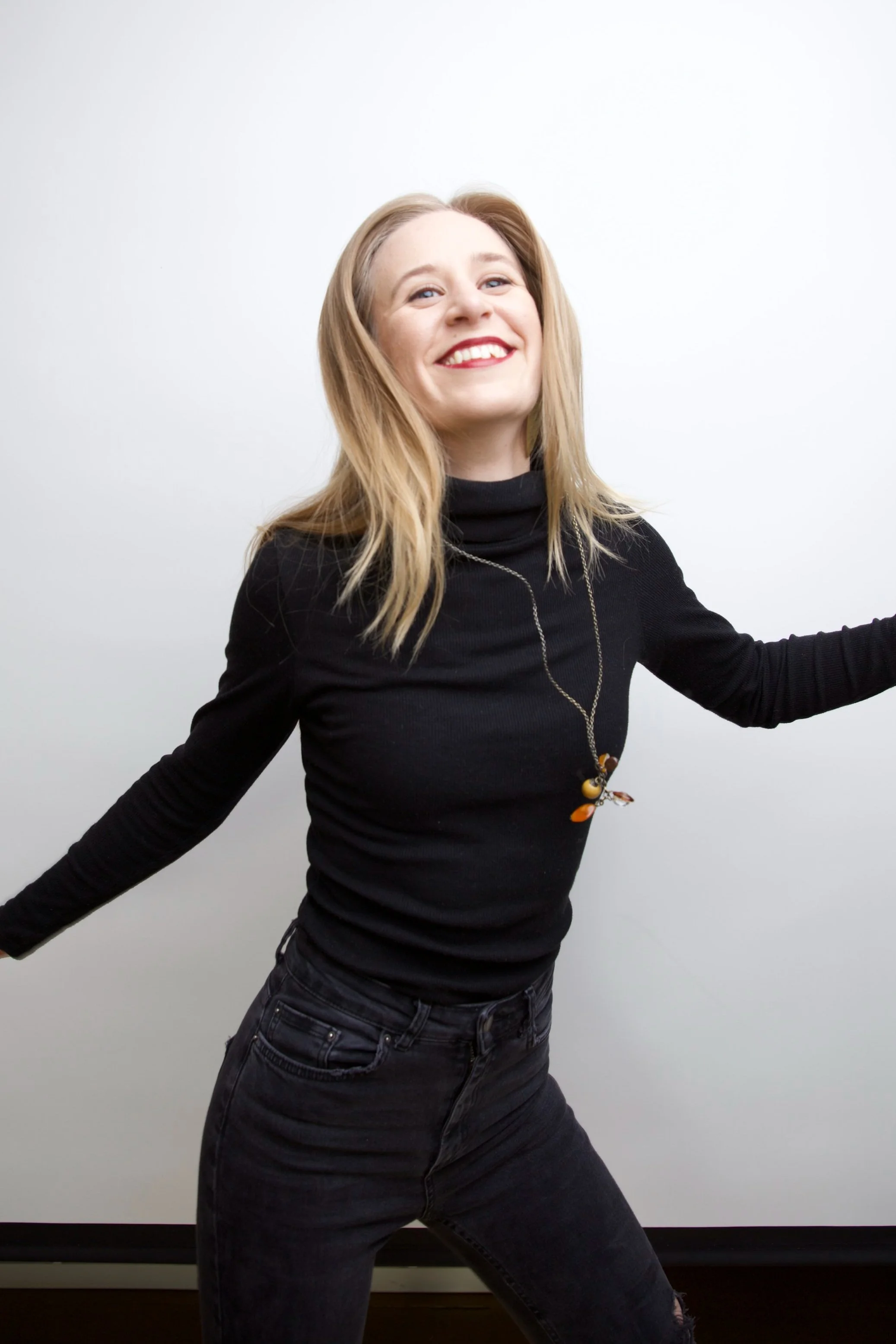 A cheerful woman with blonde hair, wearing a black long-sleeve top and black jeans, smiling with arms outstretched against a plain white wall.