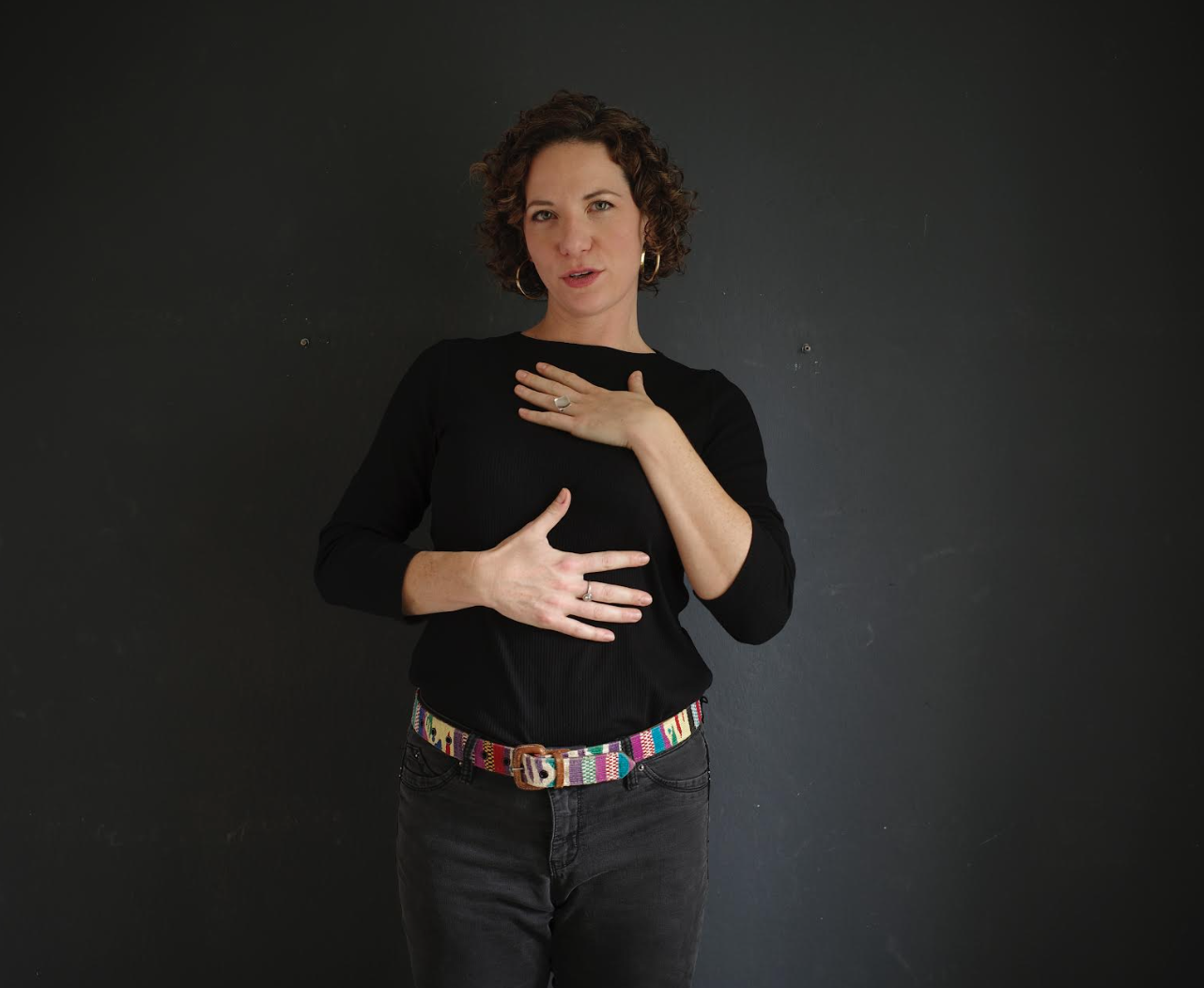 A woman with short curly hair and hoop earrings standing against a dark background, wearing a black shirt and dark pants with a colorful belt, holding her hand on her chest and stomach.