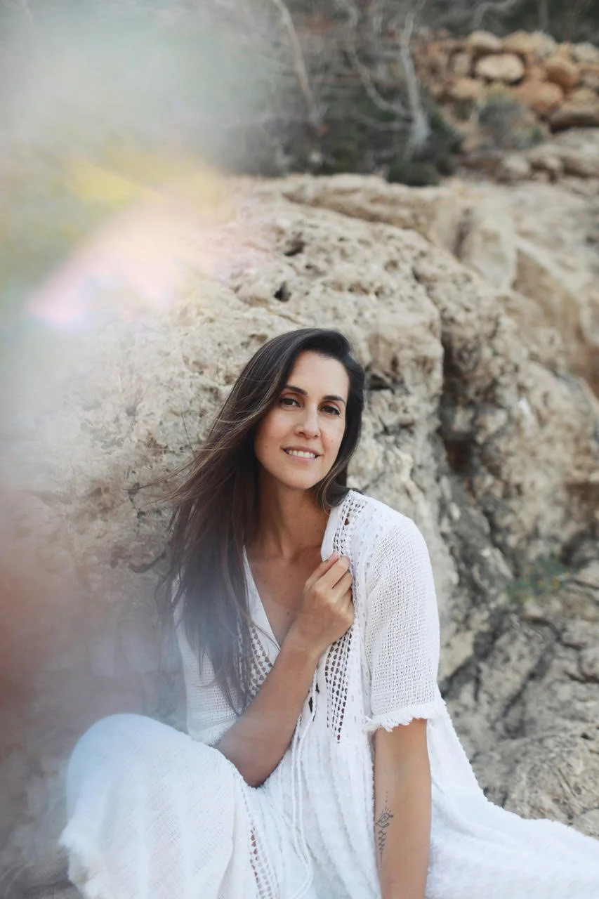 A woman with long dark hair, wearing a white crocheted dress, sitting outdoors in front of a rocky background, smiling at the camera.