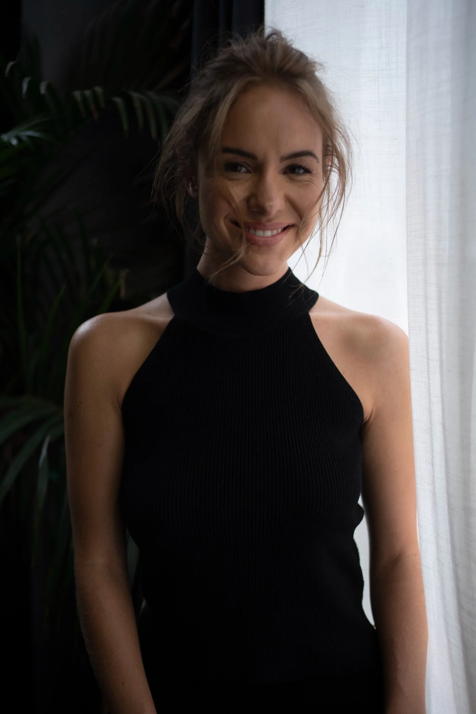 A woman smiling, wearing a black halter top, standing next to a window with white curtains and green plants in the background.