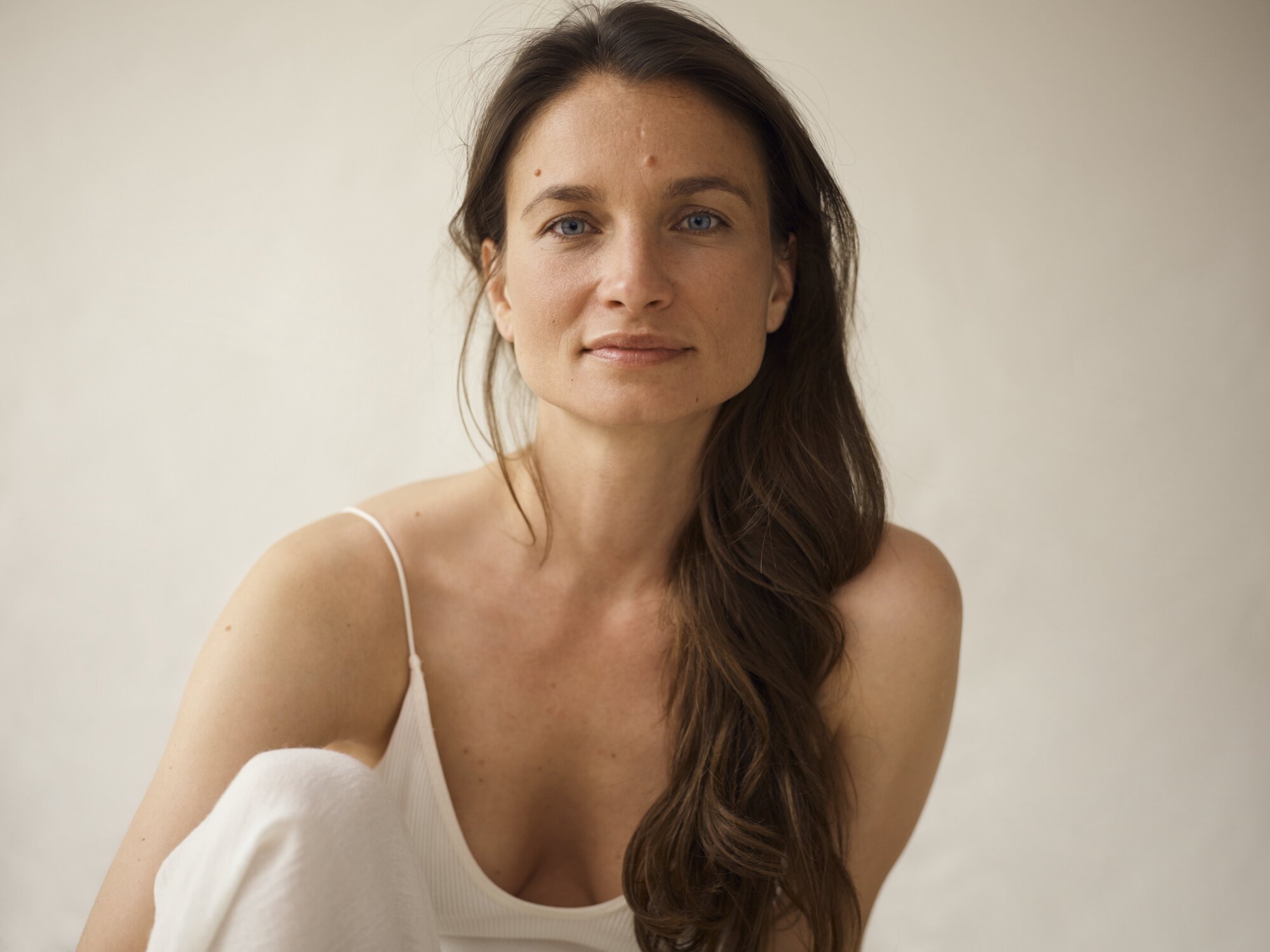 A woman with long brown hair, blue eyes, and light skin wearing a white tank top, smiling softly, against a neutral background.