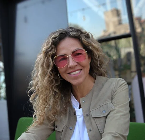 Woman with curly blonde hair wearing pink sunglasses and a beige jacket, smiling outdoors with modern glass building in the background.