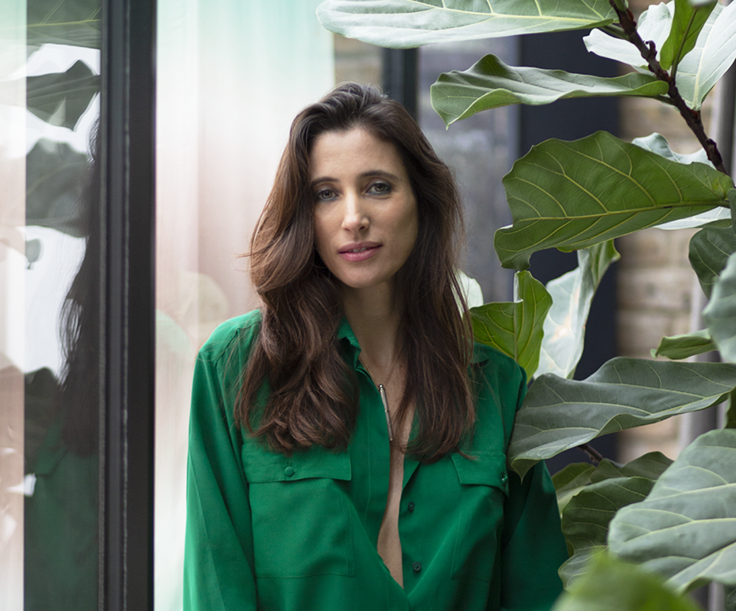 A woman with long brown hair wearing a green shirt standing among large green leaves of a plant inside a room with glass windows.