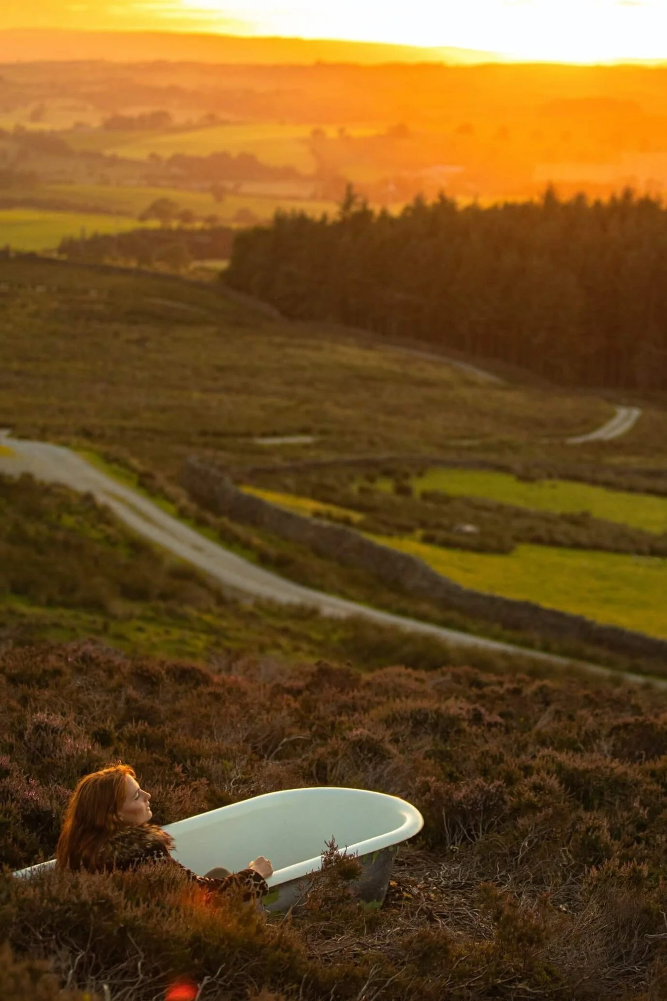 A woman with red hair in a leopard print coat sitting on the ground with a vintage white bathtub in a scenic landscape at sunset, rolling hills, and a winding road in the background.