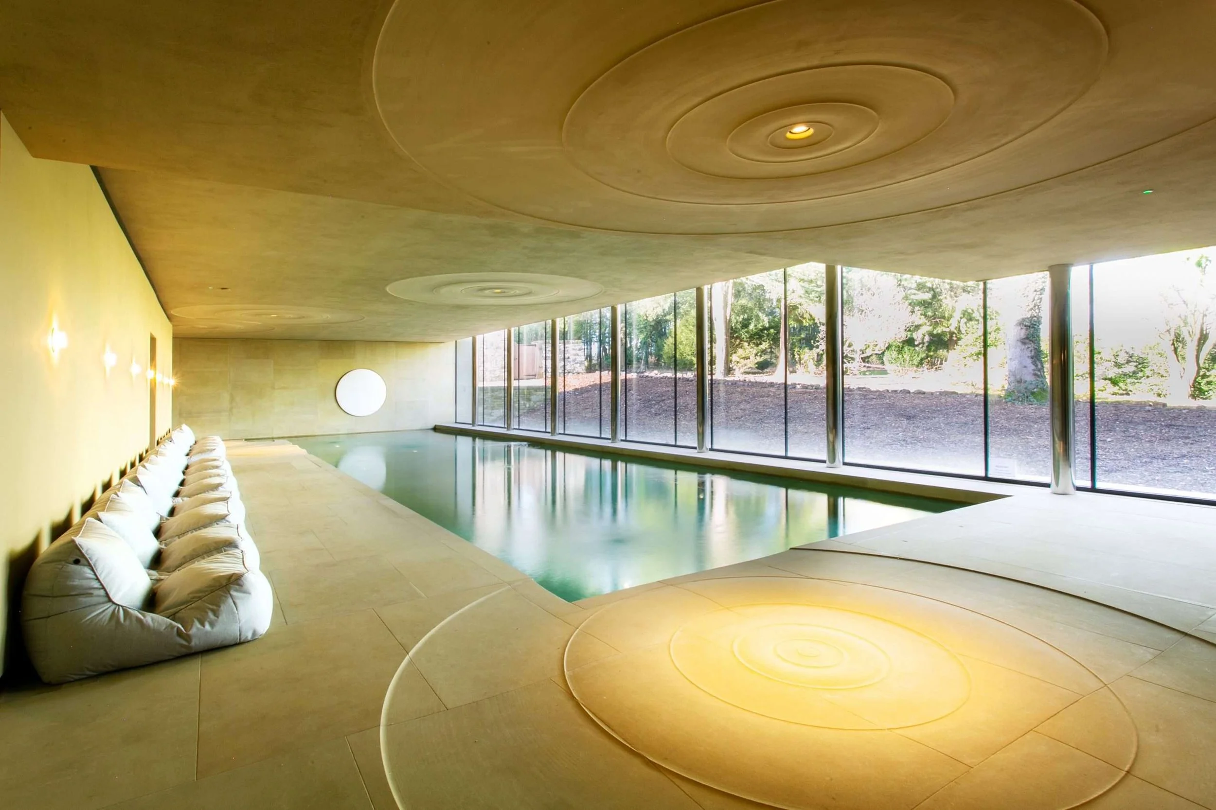 Modern indoor swimming pool area with large floor-to-ceiling windows showing outdoor trees, featuring a yellow wall with wall-mounted lights, a curved beige ceiling with circular patterns, and a row of white bean bag chairs along the wall.