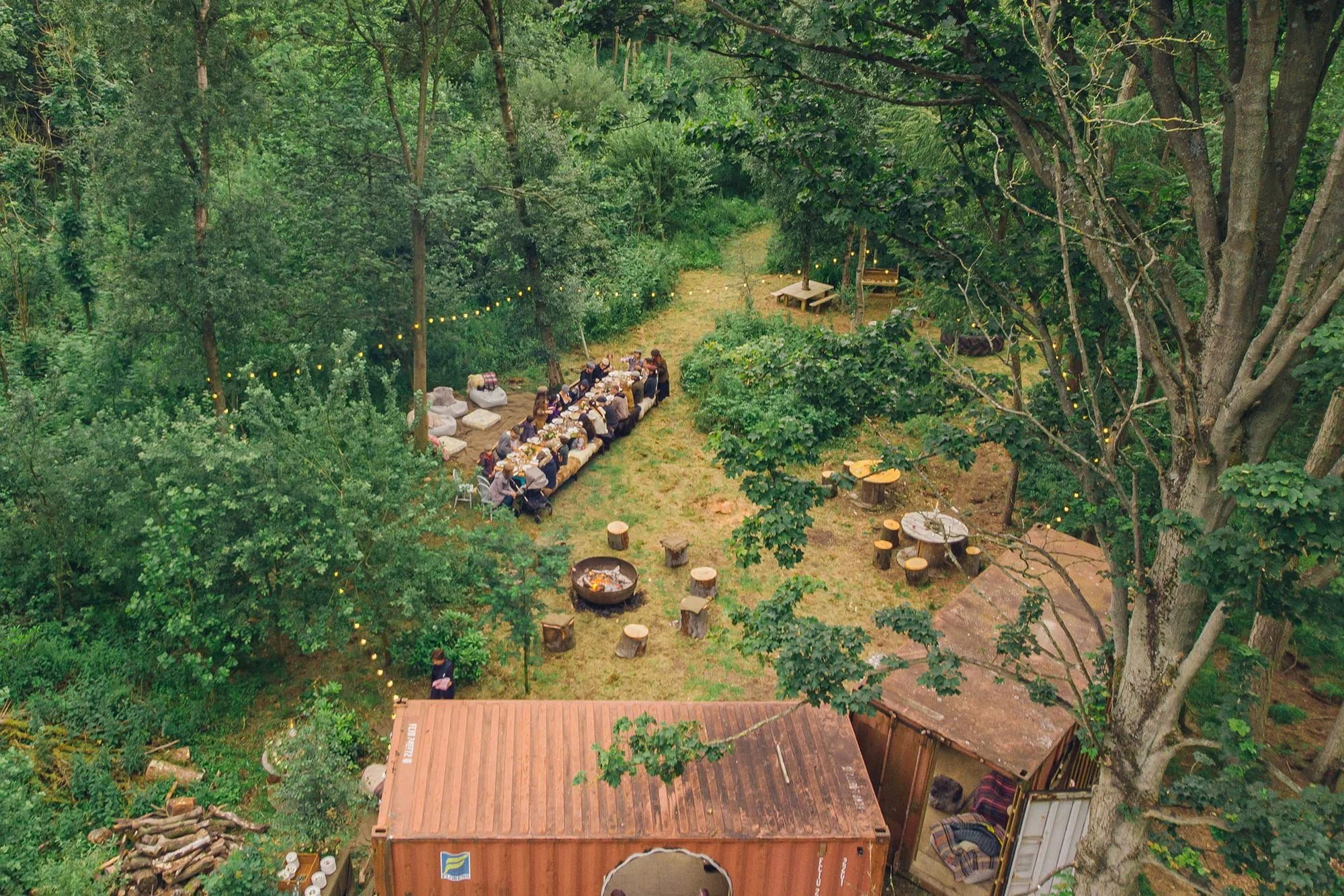 A large group of people seated at a long outdoor table in a forest setting, enjoying a meal amidst trees with string lights overhead. There are wooden logs and chairs, a fire pit, and a small wooden building nearby.