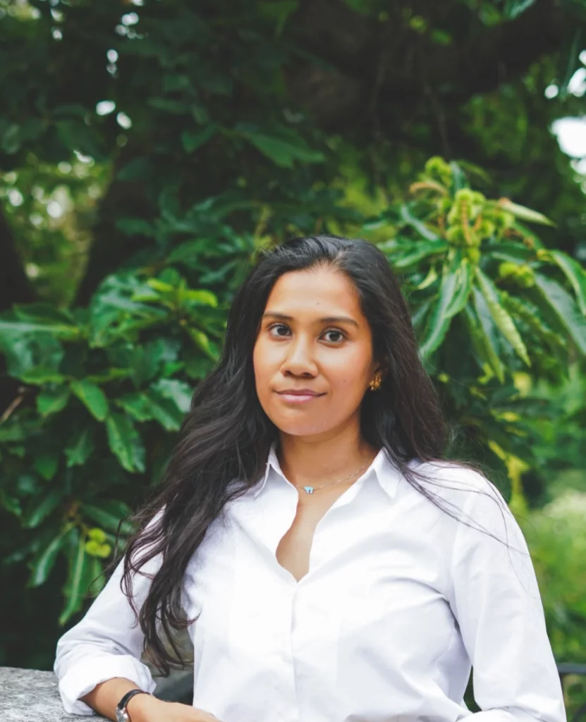 A young woman with long black hair wearing a white blouse, standing outdoors in front of green foliage.