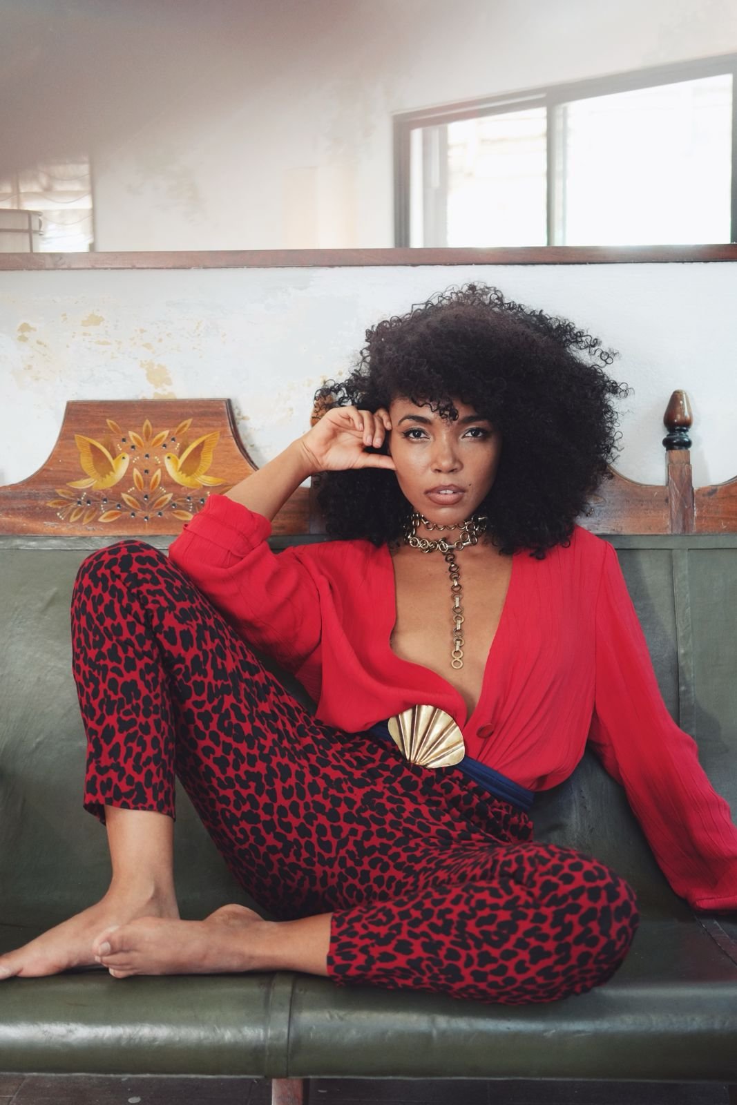 A woman with curly black hair is sitting on a green leather bench, wearing a red blouse, leopard print pants, and gold jewelry, posing with her head resting on her hand in front of a wooden headboard and a window.