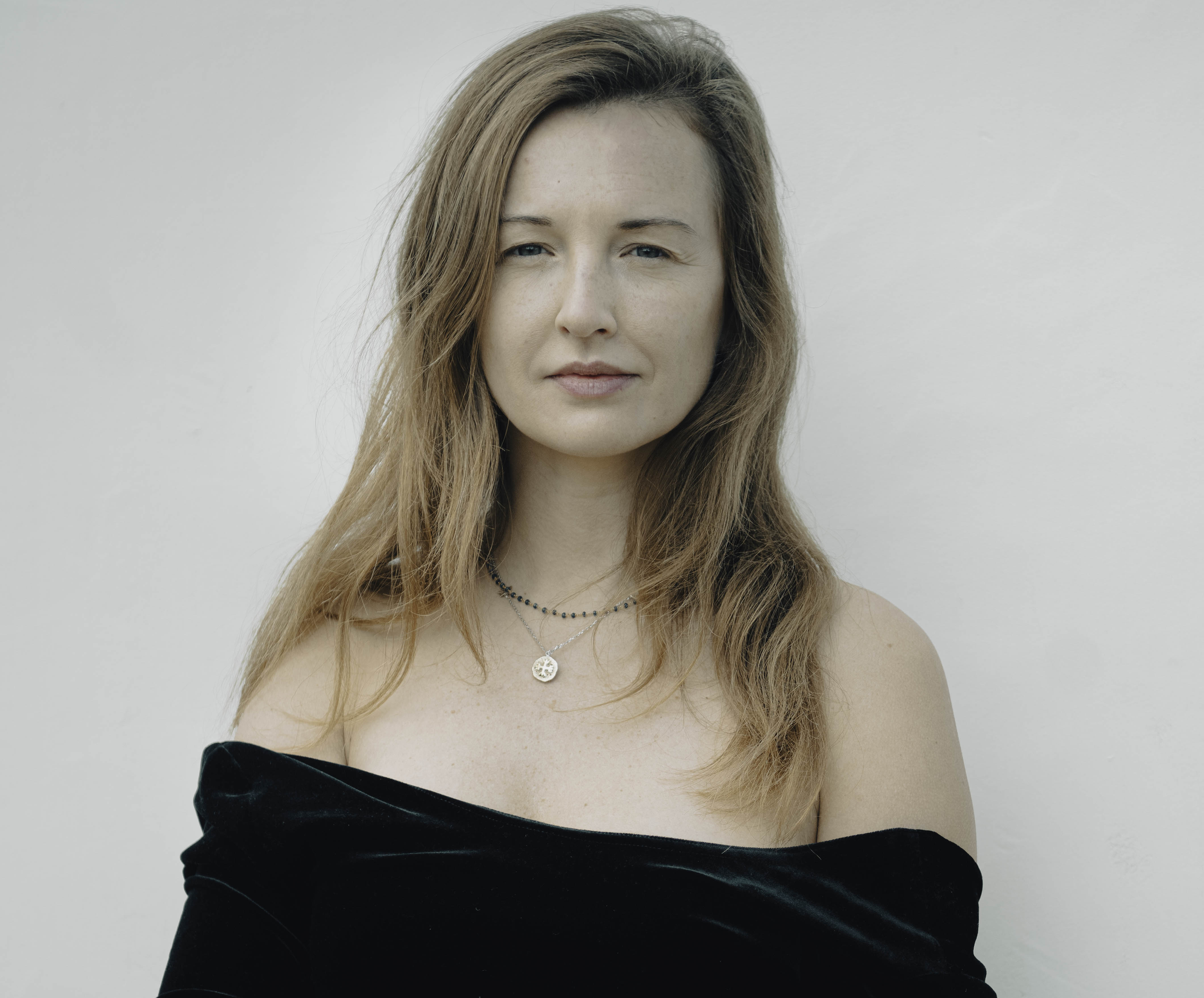 A woman with long, wavy, light brown hair wearing a black off-the-shoulder top and layered necklaces, standing against a plain light-colored background.