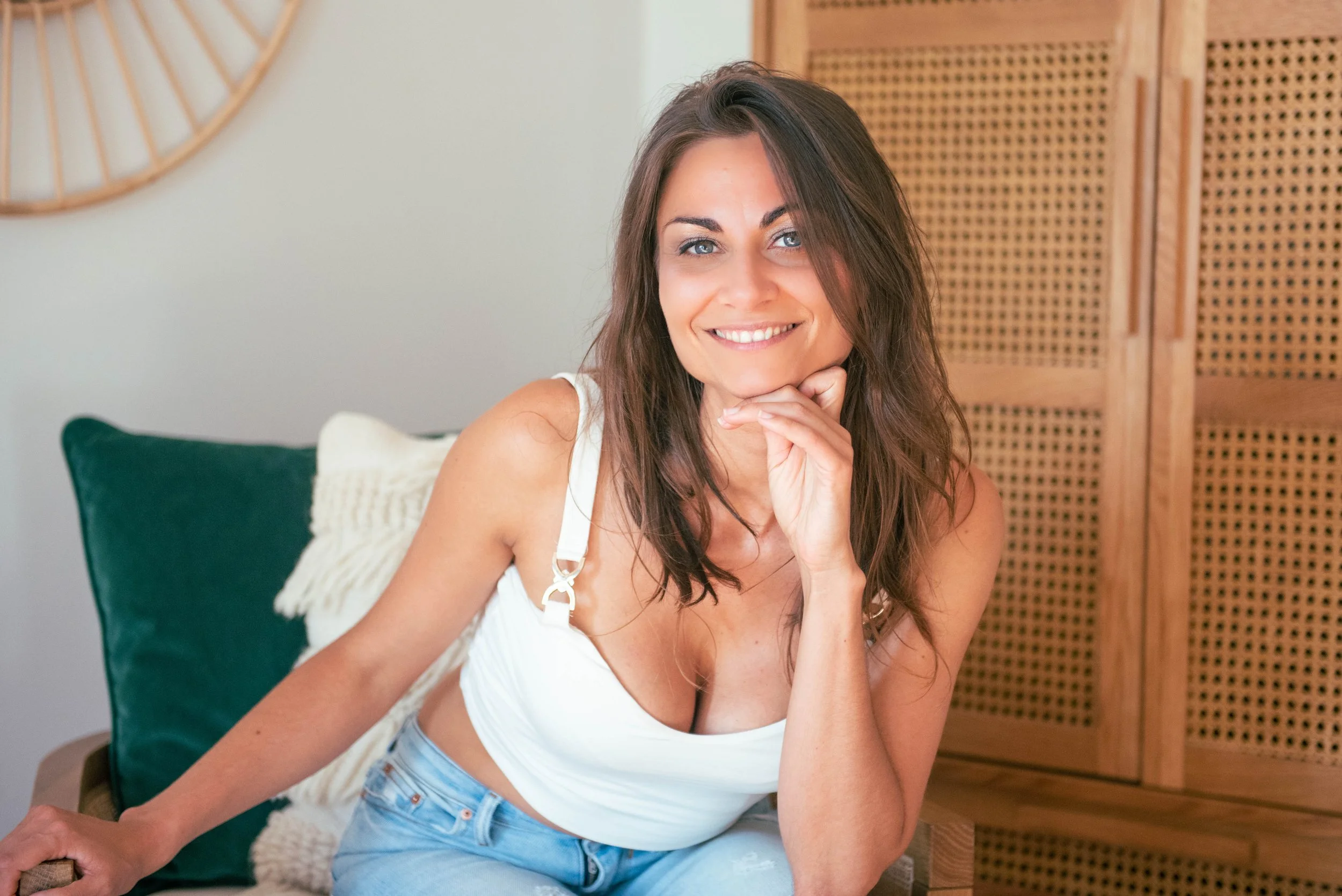 A woman with long brown hair wearing a white tank top and light blue jeans, smiling while sitting on a green cushion with a white pillow behind her, in a room with a wooden partition and a wall clock in the background.