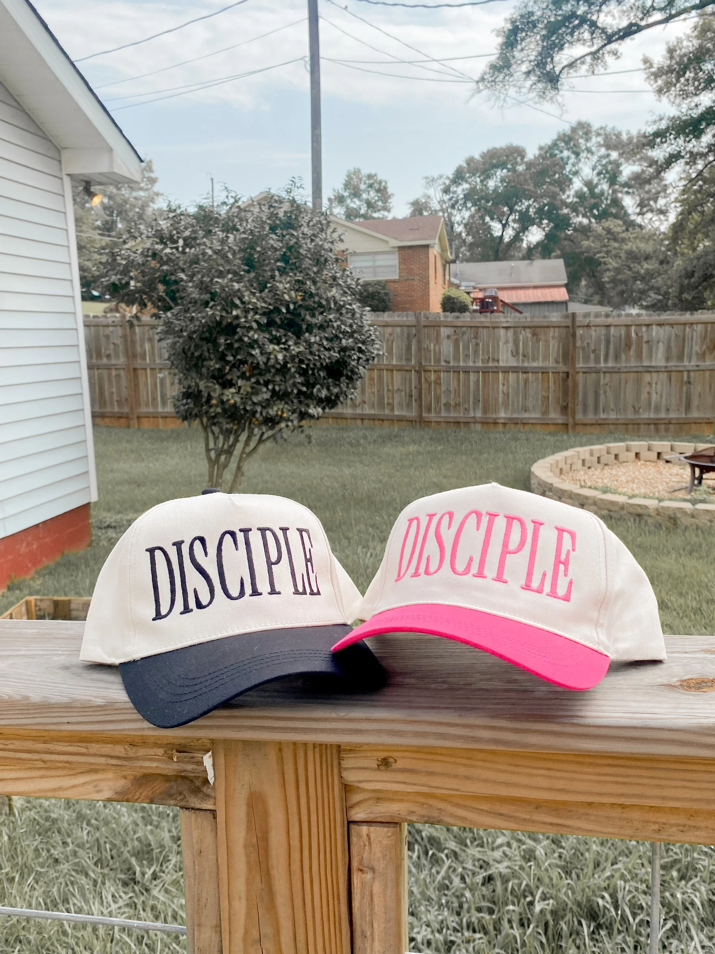 Three caps on a wooden table, two with the word 'DISCIPLE' embroidered on the front, one with pink and white coloring.