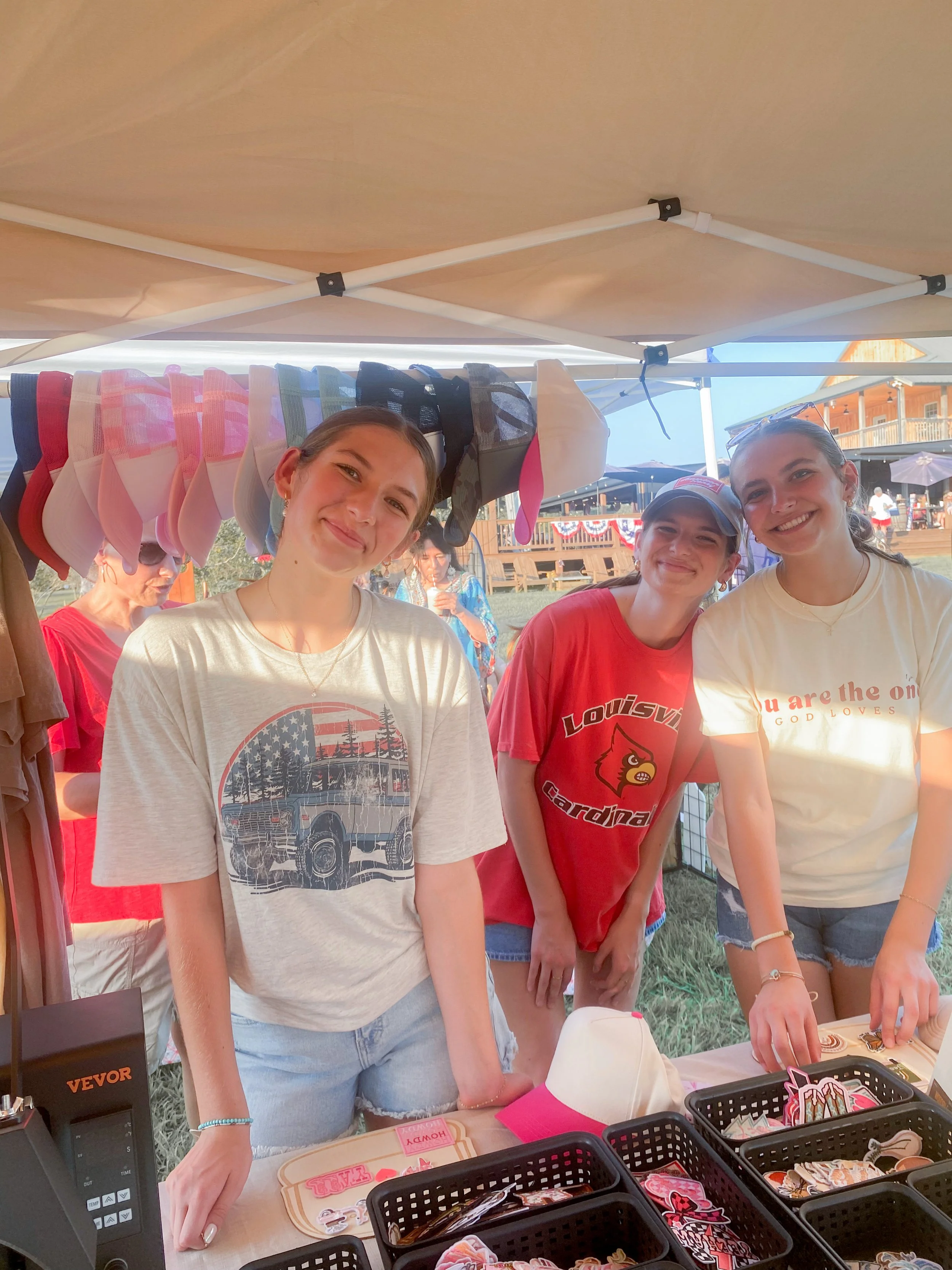Three young women smiling at a table with various stickers and merchandise at an outdoor event under a canopy.