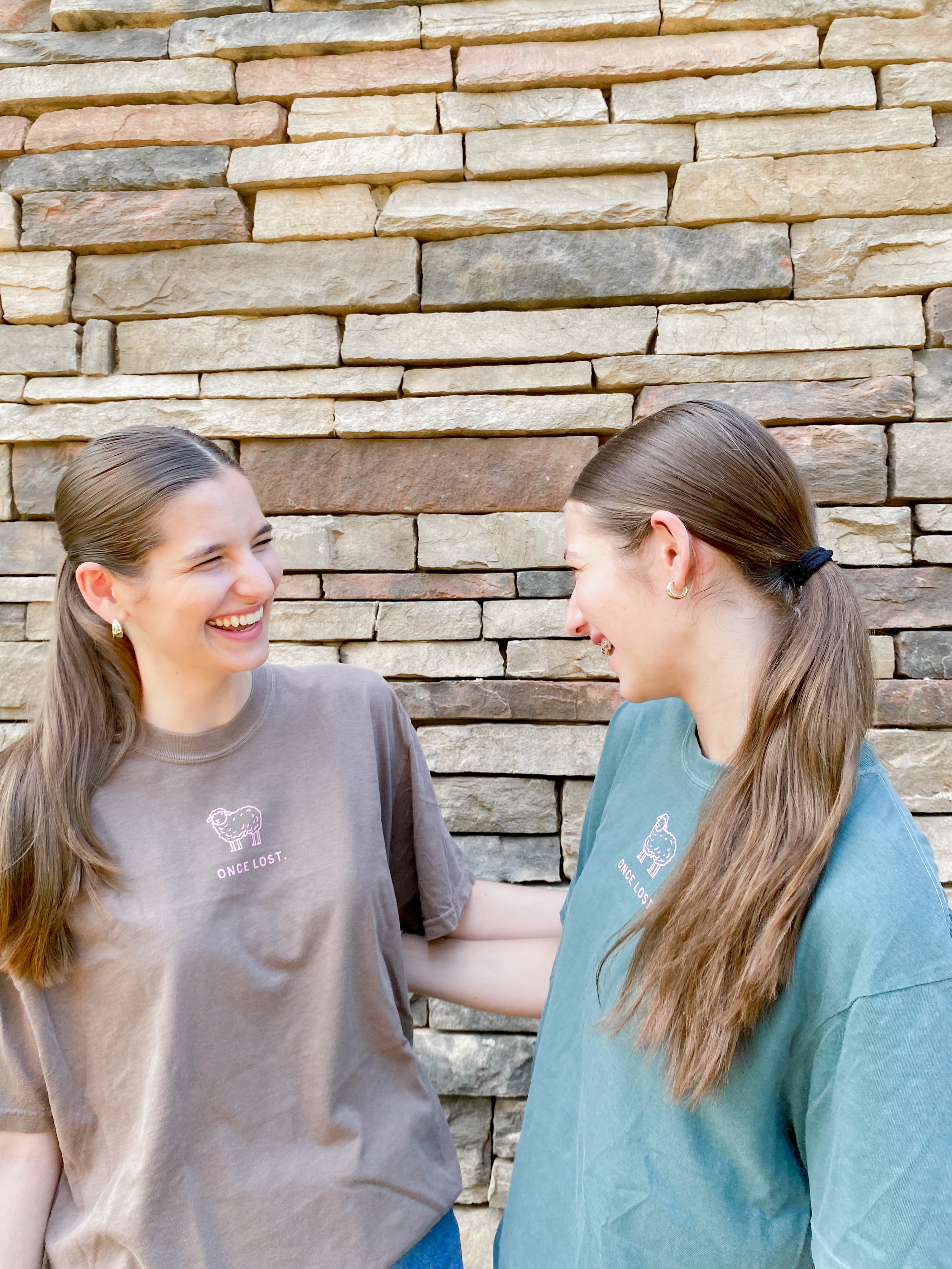 Two young women with long hair and gold hoop earrings are smiling and laughing while looking at each other against a stone wall. They are wearing casual t-shirts with a sheep and the text "ONCE LOST." on them.