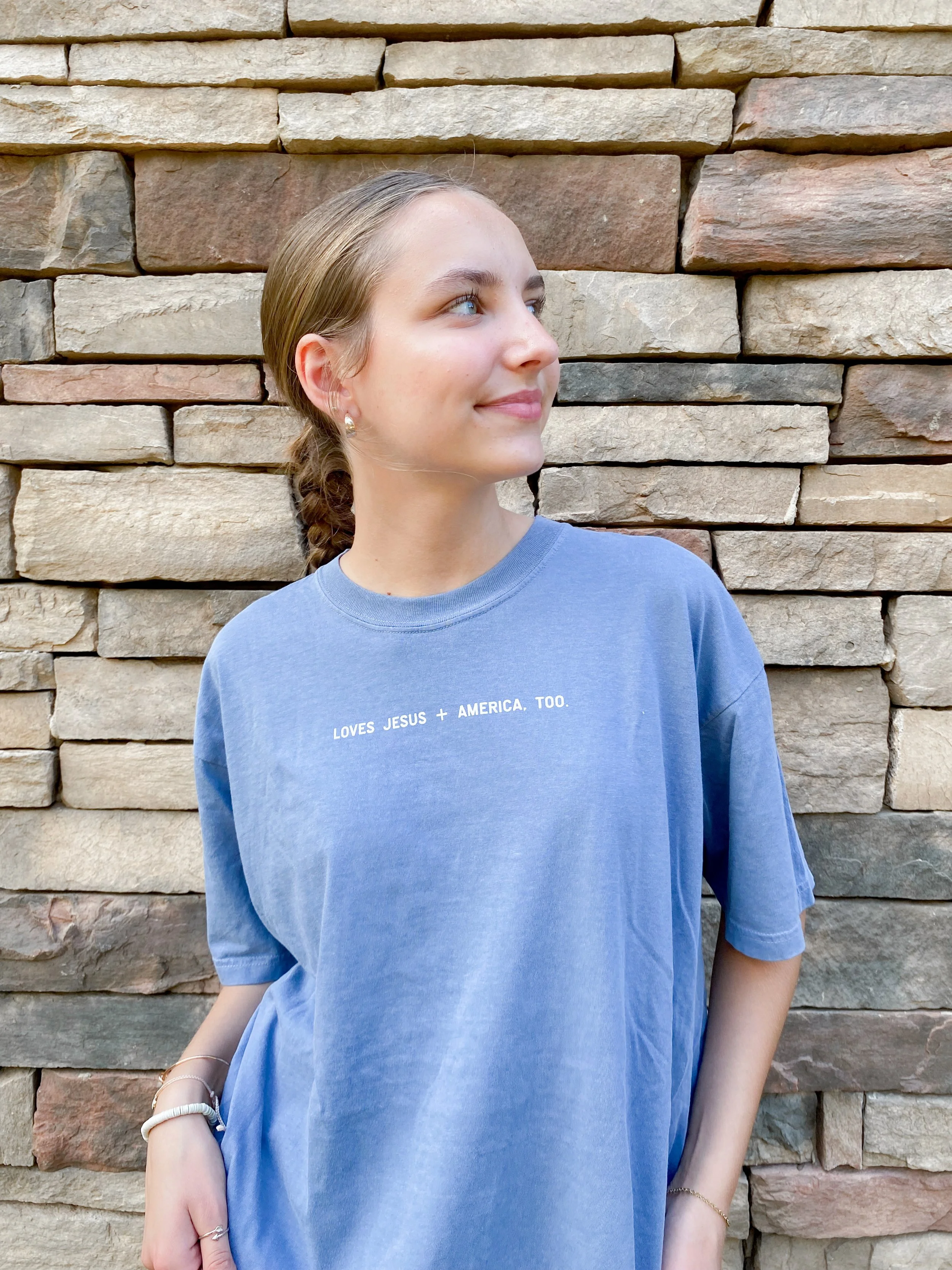 A young woman with blonde hair pulled back in a braid, wearing a blue T-shirt with the text 'LOVES JESUS + AMERICA, TOO.' and minimal jewelry, posing against a stone wall.