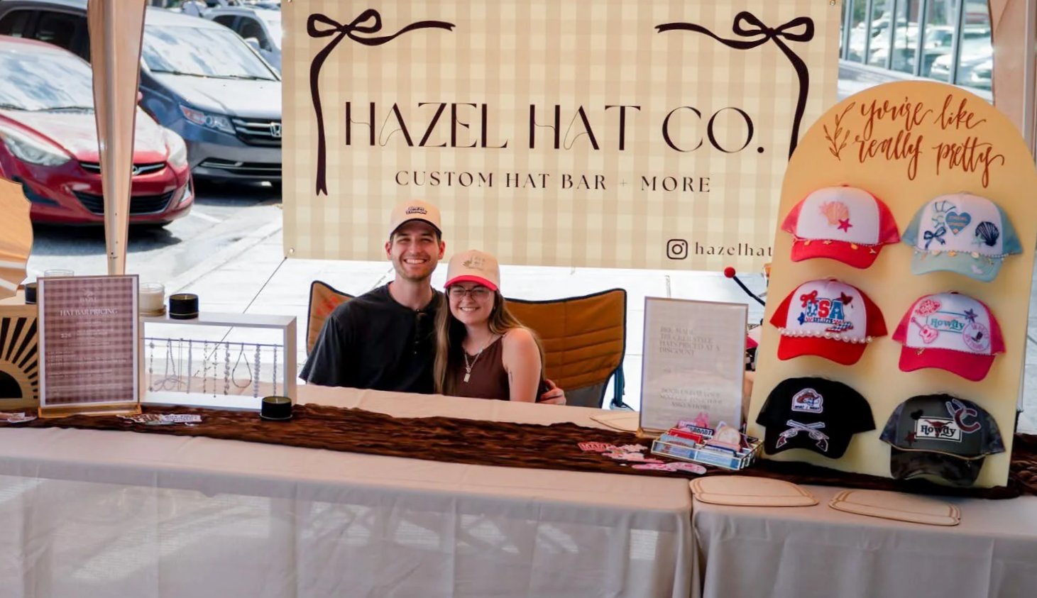 A smiling man and woman wearing hats sitting behind a trade booth for Hazel Hat Co at a market or fair. The booth displays custom hats and accessories with a large sign reading 'Hazel Hat Co. Custom Hat Bar + More' and decorated with various hats and patches.