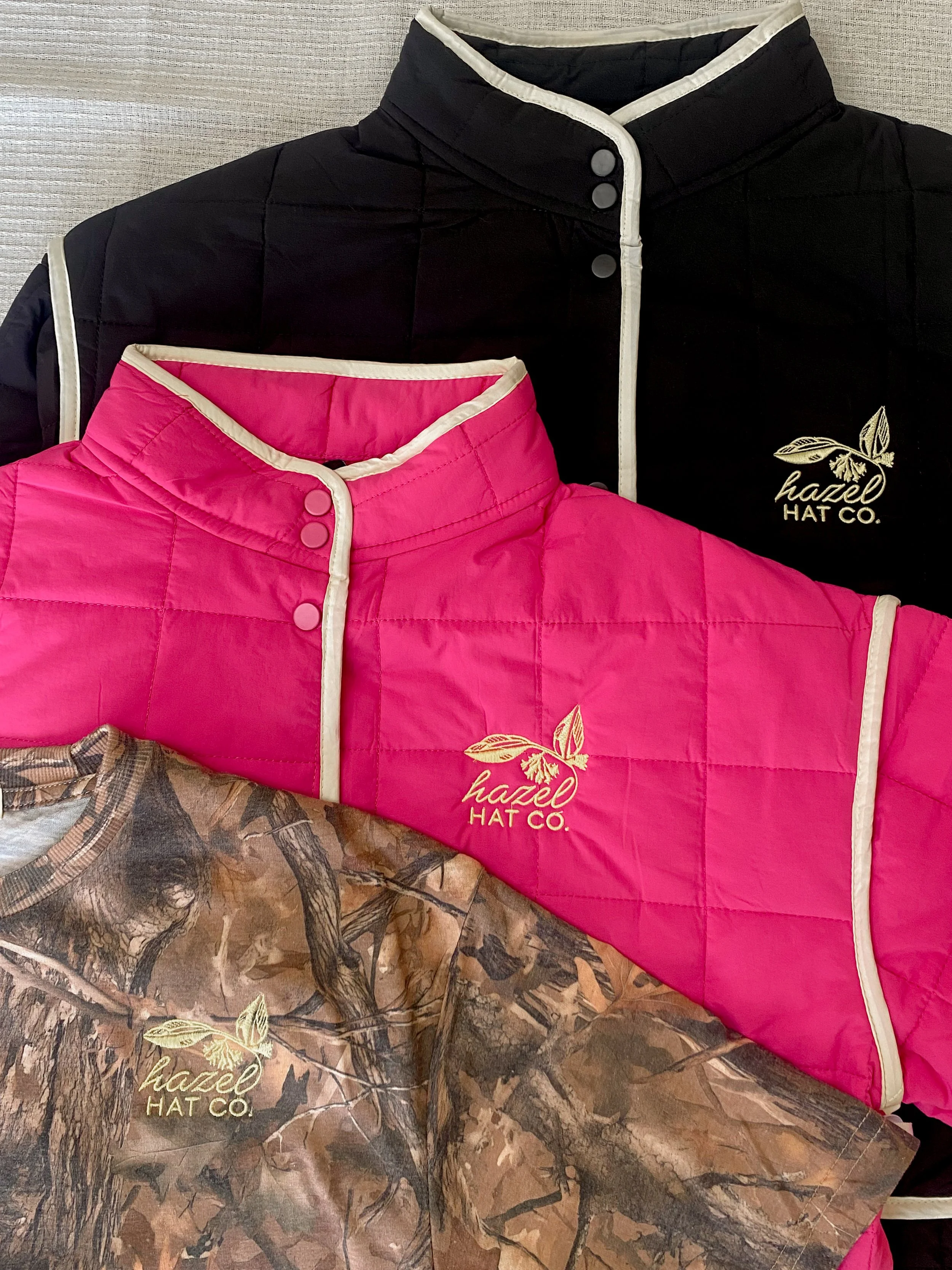 Two quilted jackets, one black and one pink, with a logo and the words "hazel HAT CO." on each, and a camouflage-patterned item underneath the pink jacket.