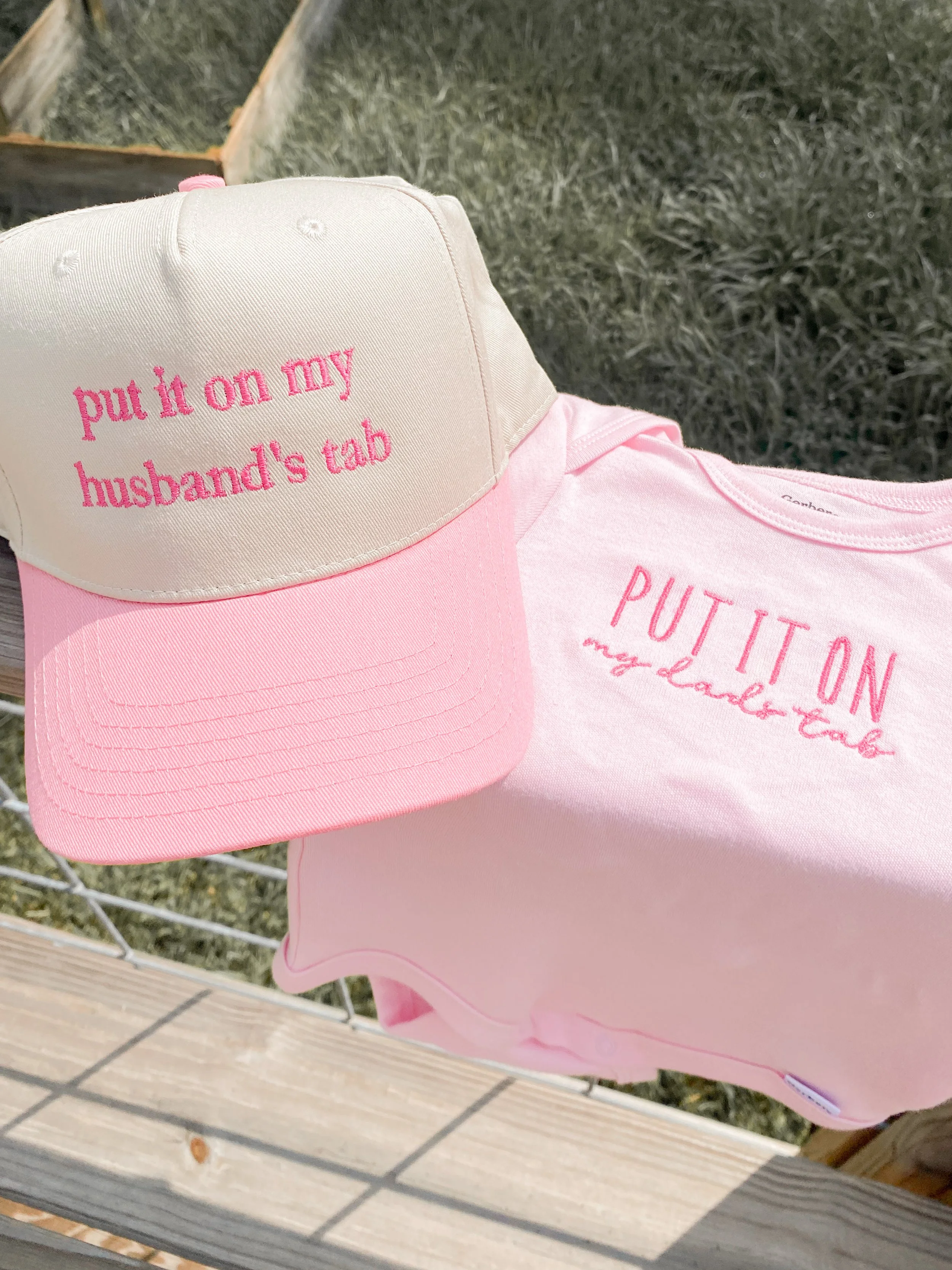 Pink and white baseball cap with embroidered text, lying on pink cloth with printed text, on wooden and grass surface