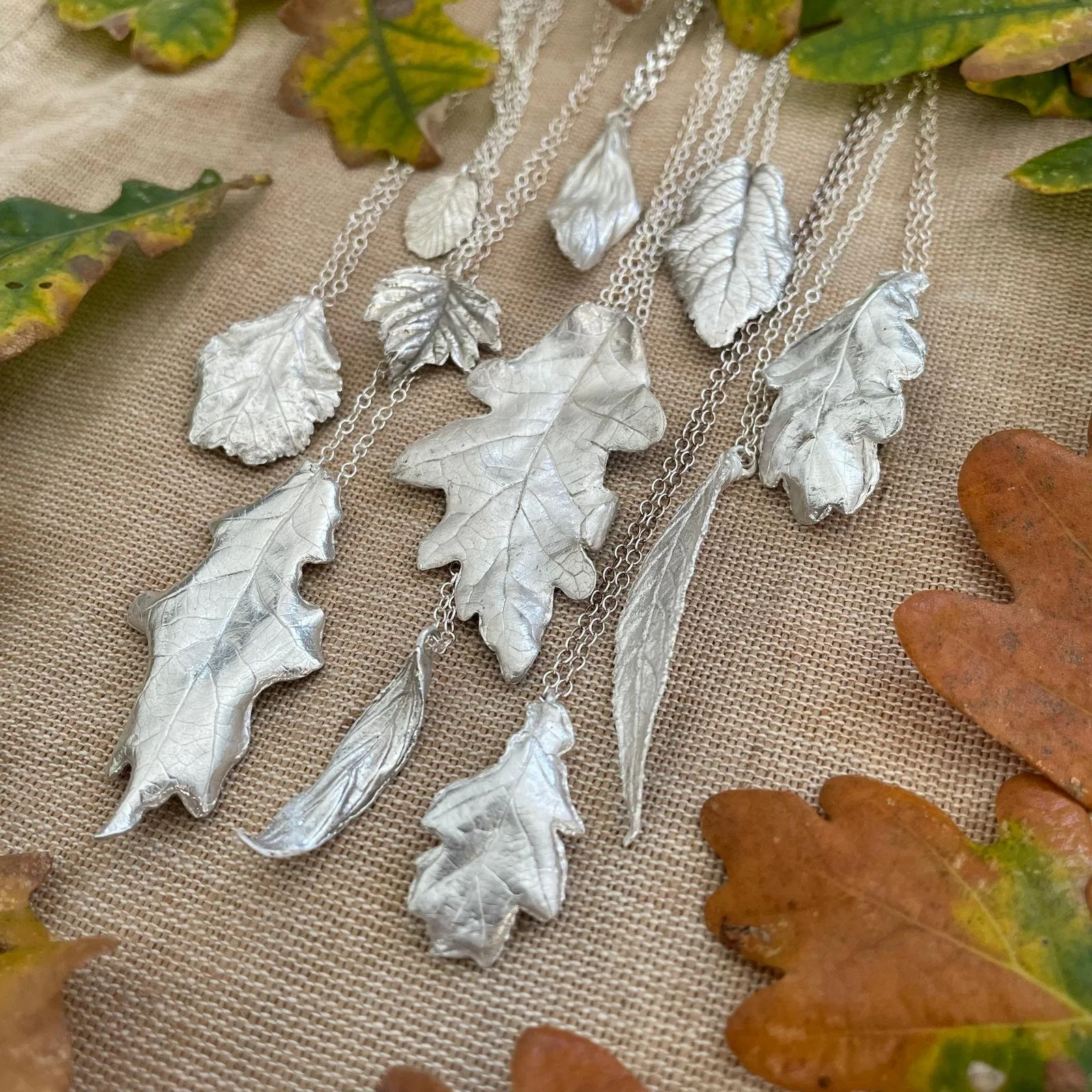 Silver leaf-shaped jewellery necklaces arranged on a textured surface with autumn leaves around them.