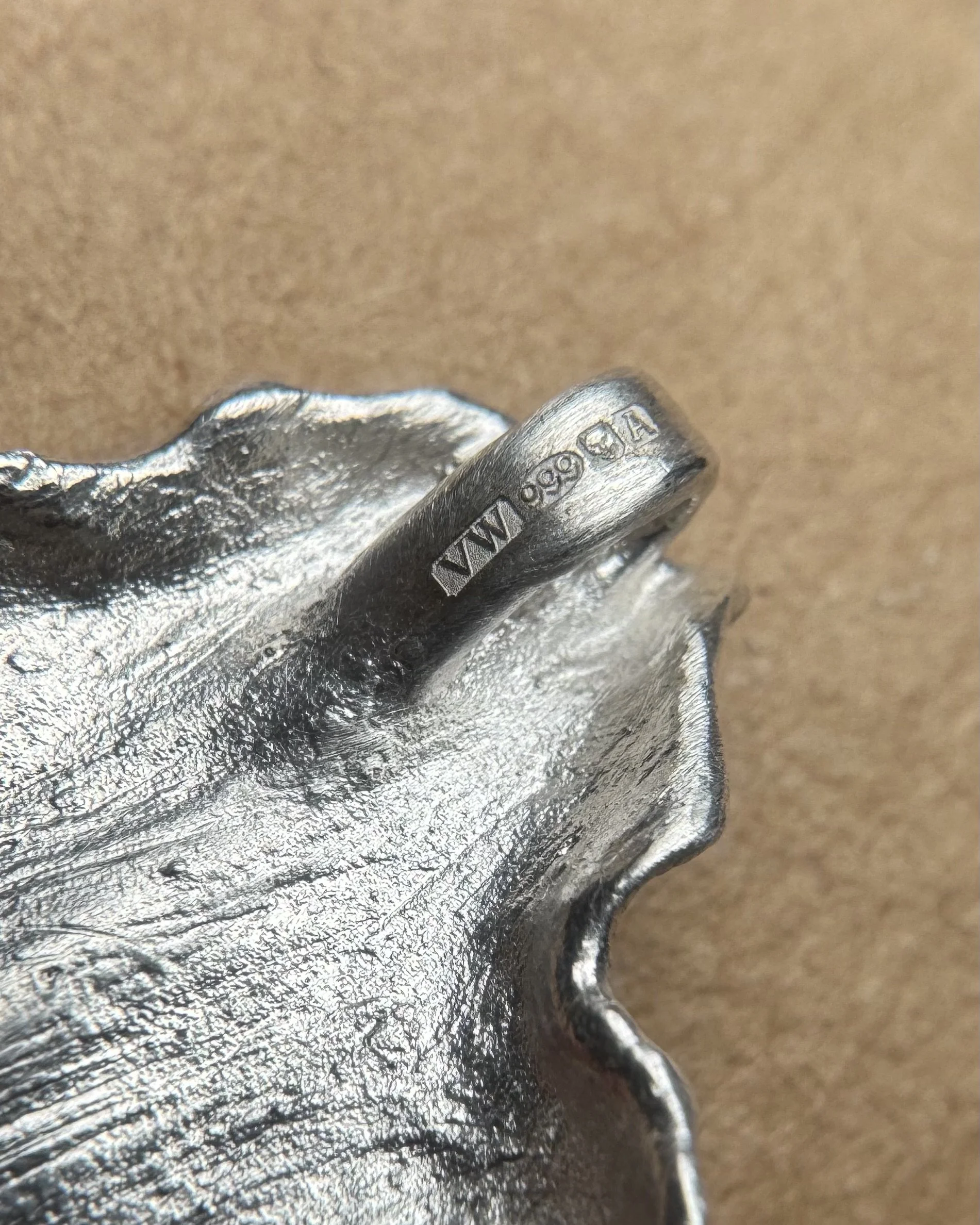 Close-up image of an irregularly shaped silver-colored metallic object with engraved markings on one edge, resting on a textured brown surface.