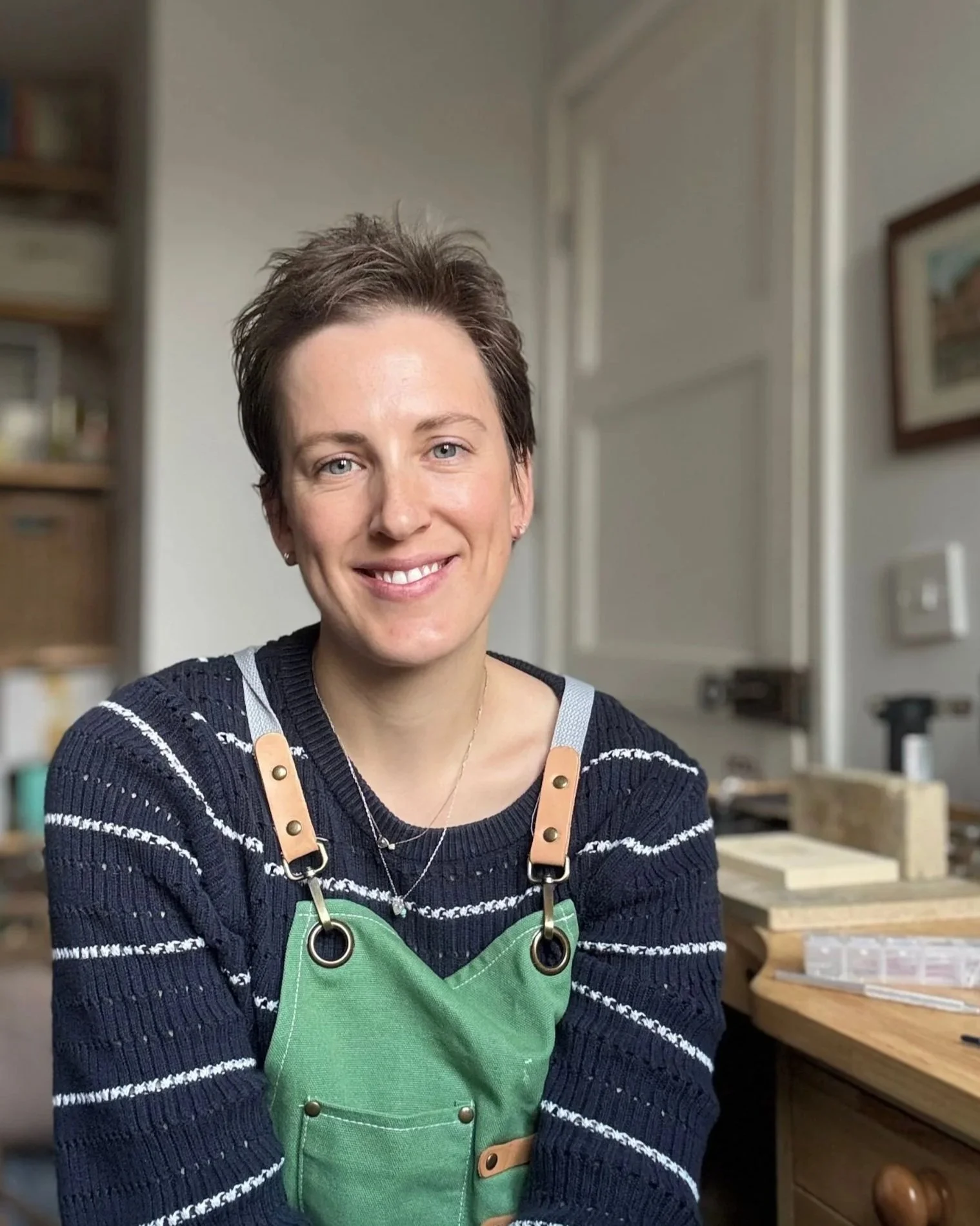 A woman with short brown hair smiling, wearing a black and white striped sweater and a green apron with leather straps, inside a woodworking or craft workshop with wooden furniture in the background.