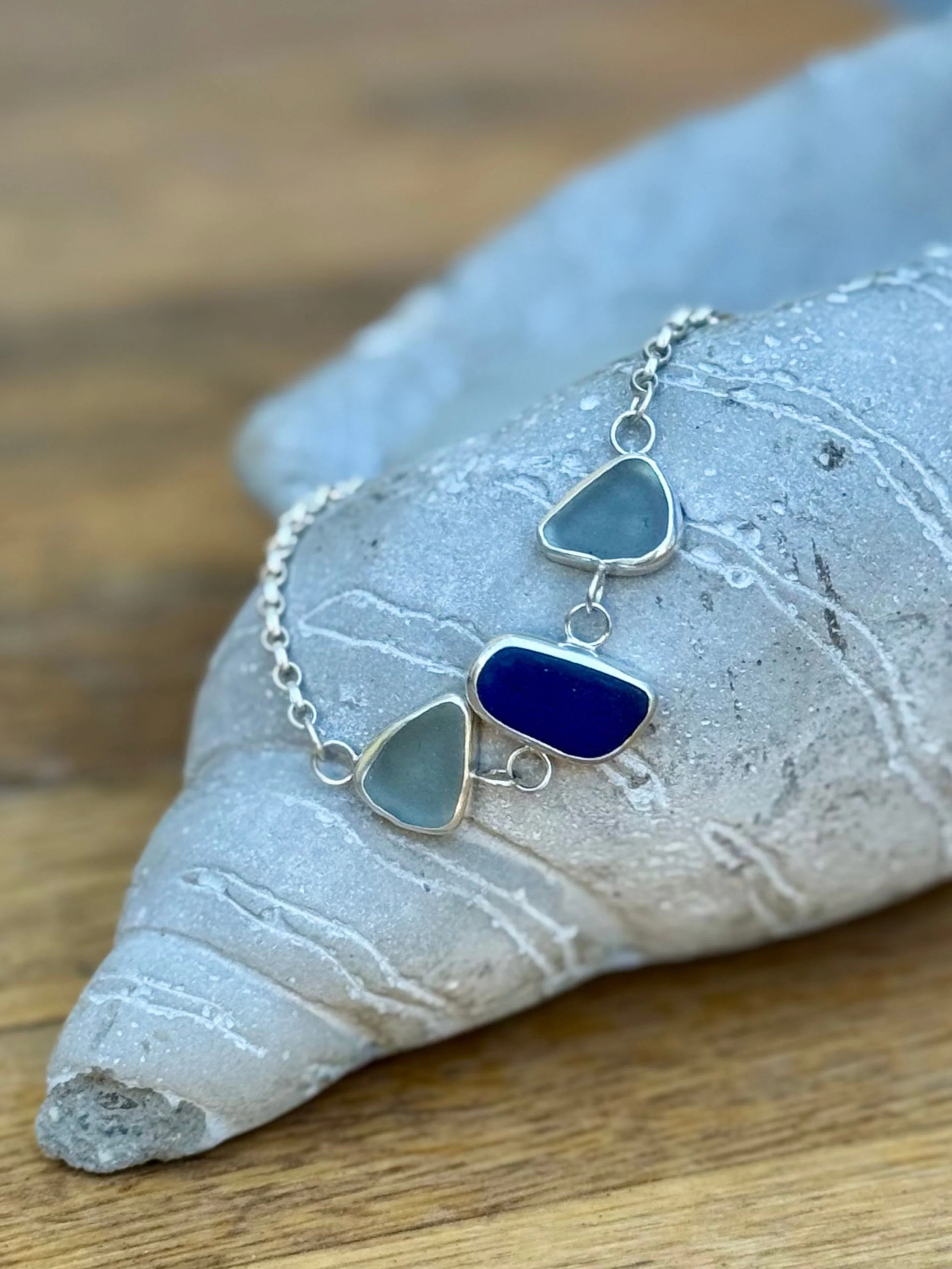 A silver necklace with six triangular and square-shaped dark blue and gray stones rests on a white, textured shell on a wooden surface.