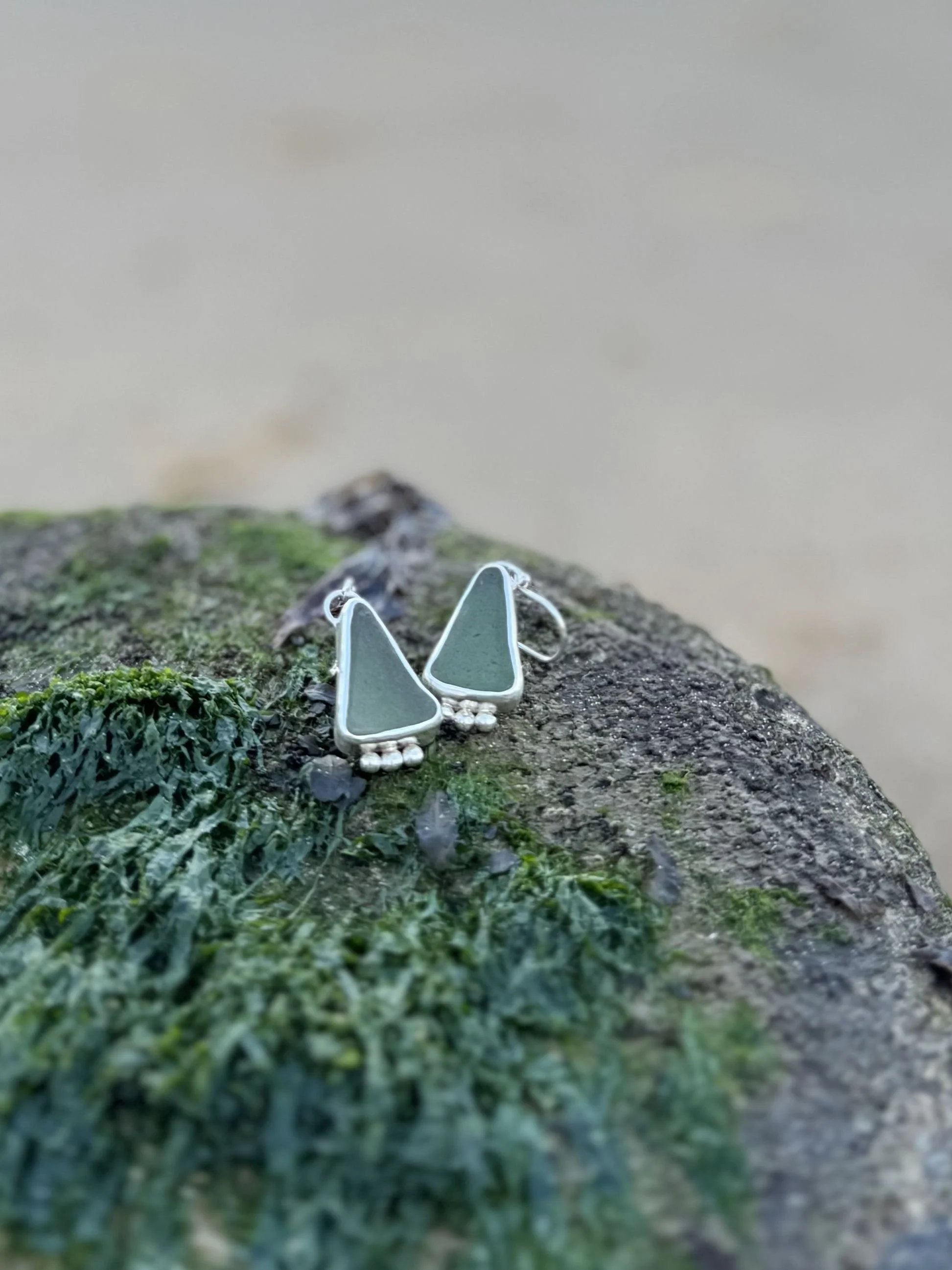 A close-up of a pair of silver earrings with green sea glass and silver accents, resting on a mossy and rocky surface near a body of water.