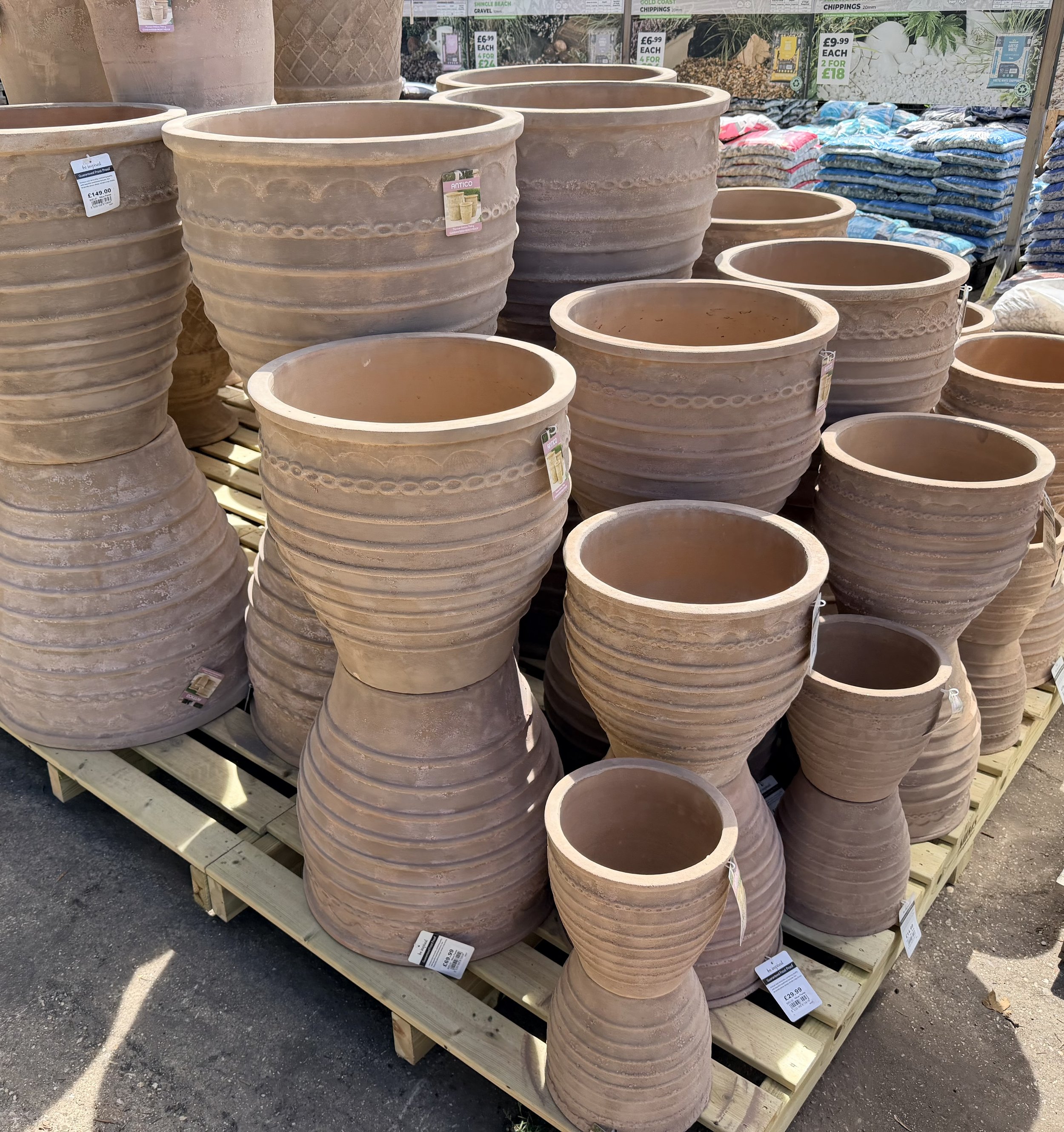 Several large terracotta plant pots with decorative banded designs on a wooden pallet outdoors at a garden center, with price tags and gardening supplies visible in the background.