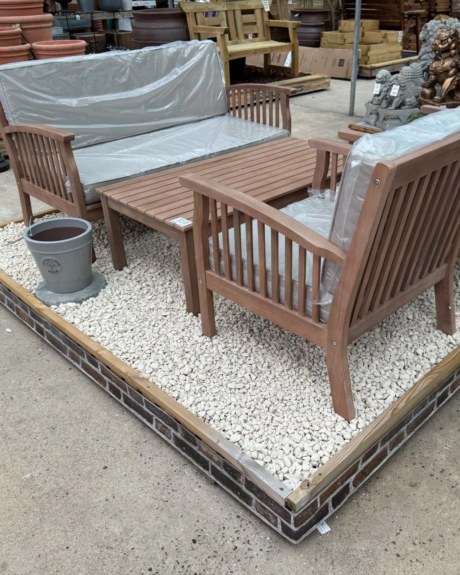 Outdoor patio furniture set consisting of a wooden slatted sofa, a matching wooden slatted armchair, and a rectangular coffee table, all covered with plastic protective wrapping. The furniture is placed on a gravel bed surrounded by a brick border, with gardening supplies and decorative statues visible in the background.