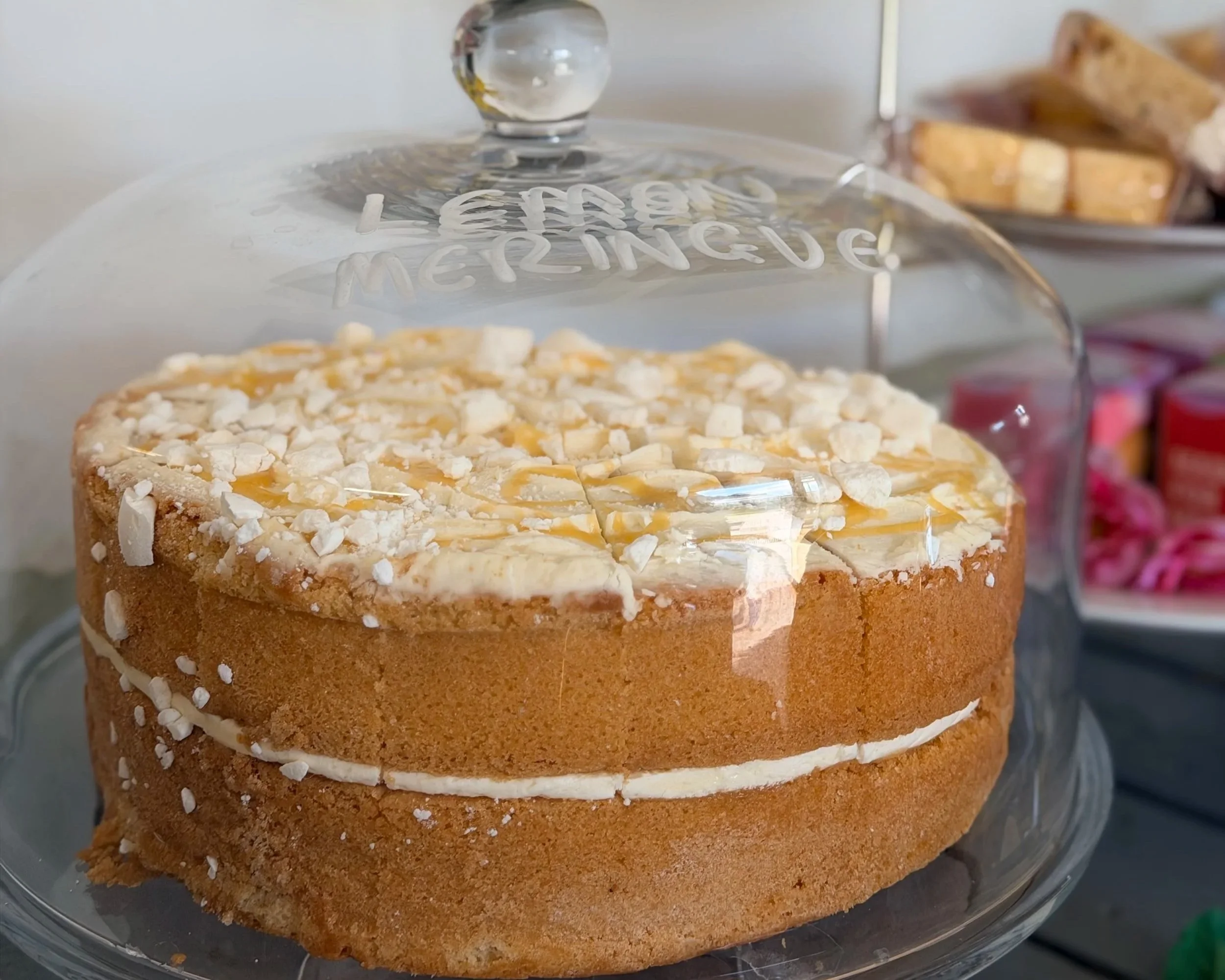A layered dessert cake with white frosting, topped with crushed white candy pieces, behind a glass cake dome. The dome has a knob at the top and the word 'MERINGUE' written on it.