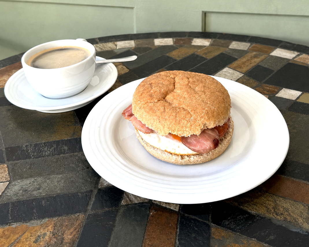 Breakfast consisting of a bagel sandwich with egg, ham, and tomato on a white plate, accompanied by a cup of coffee with a spoon on a saucer.