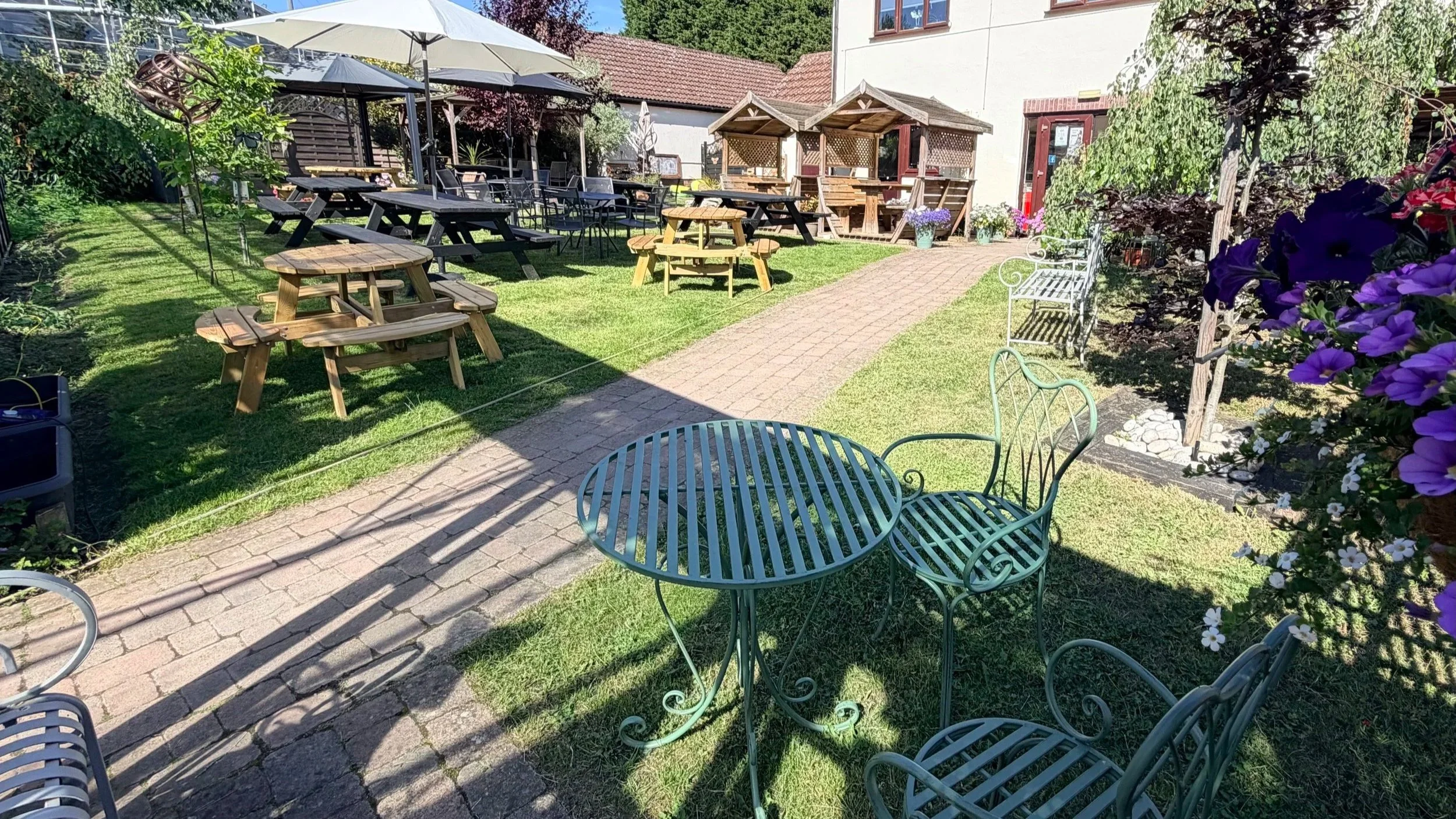 A sunny outdoor patio with green metal tables and chairs, a brick pathway, potted flowers, parasols, and shaded sitting areas surrounded by greenery and a building with a red-tiled roof.