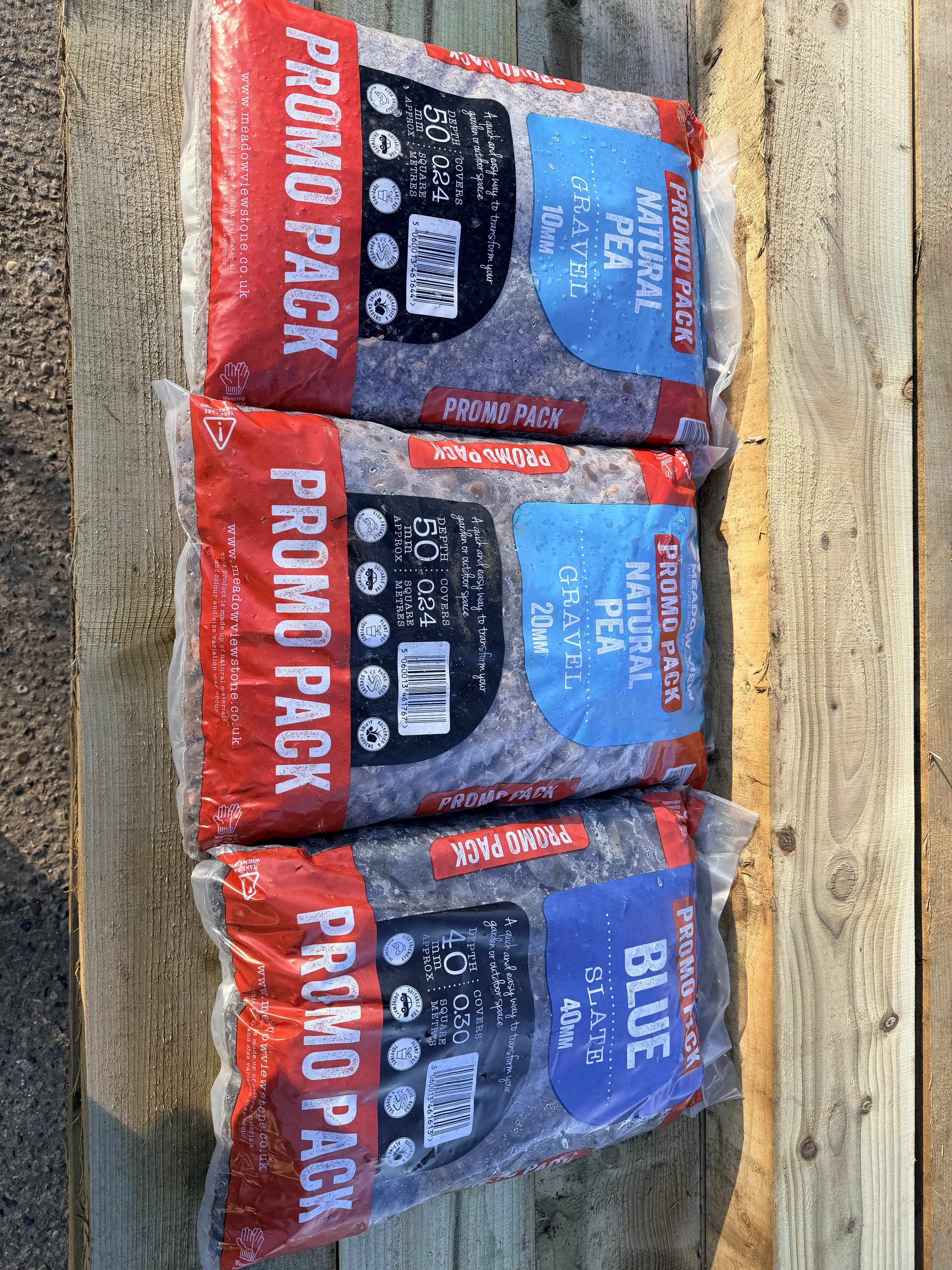 Two bags of natural pea gravel 10mm and 20mm in sealed plastic packaging, placed on a wooden surface outdoors.