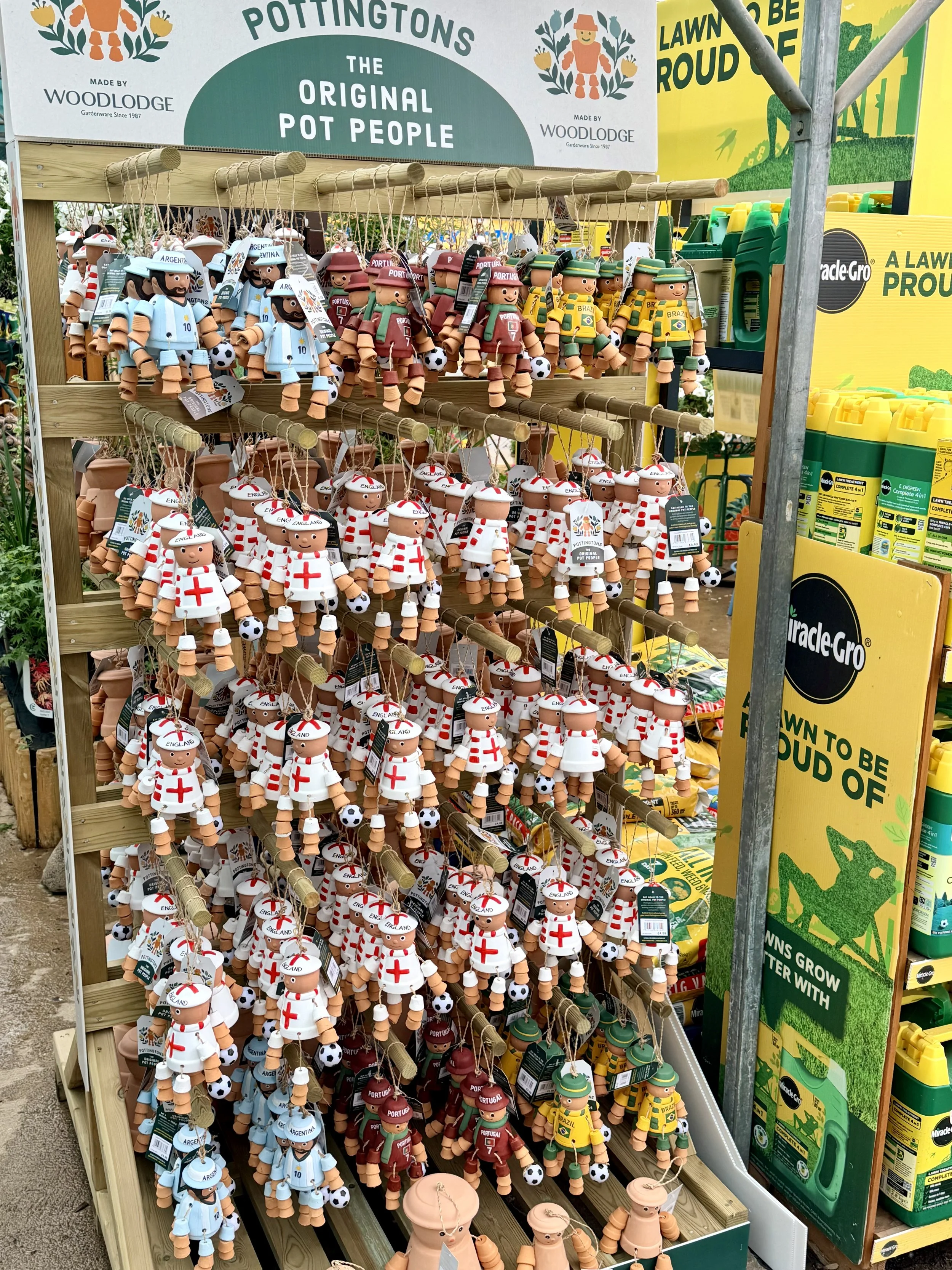 Display of pot-shaped figurines representing soccer players and national teams, organized on wooden shelves at a garden store section. The figurines depict players in team uniforms with small soccer balls, labeled with country names such as Argentina