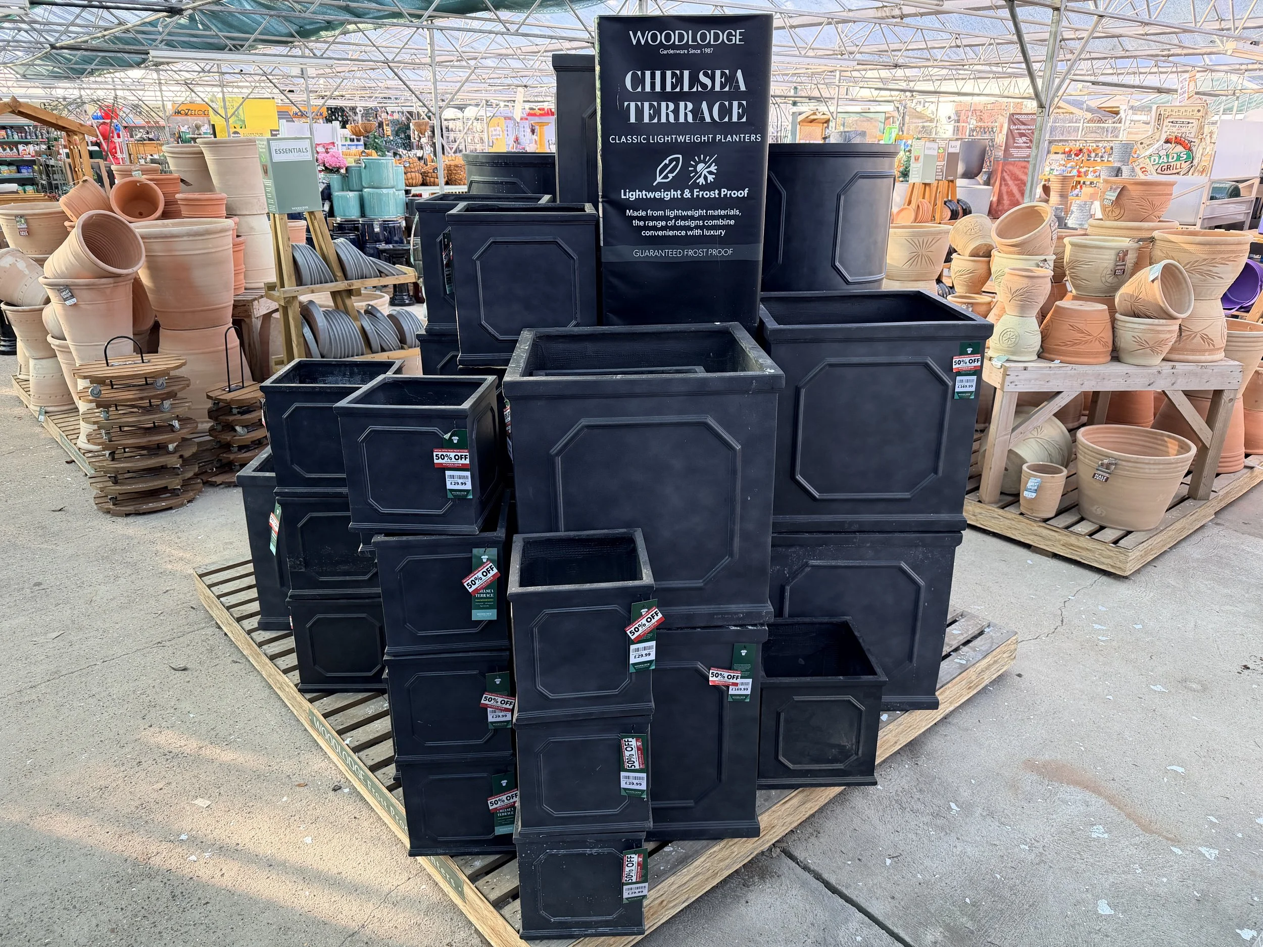 Display of black plastic planters in different sizes at a garden center, with a sign reading 'Chelsea Terrace Classic Lightweight Planters' and descriptions of the planters being lightweight, frost proof, and made from lightweight materials.
