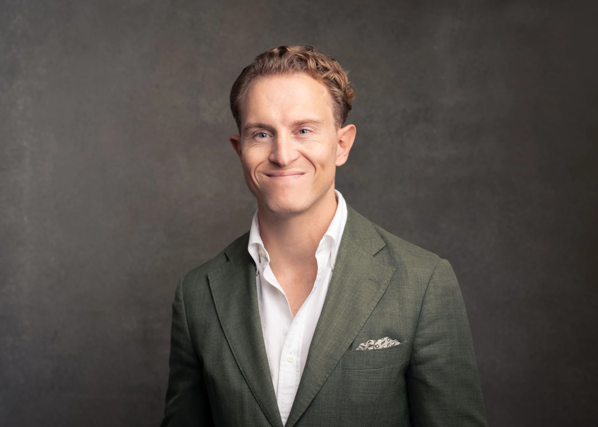 A man in a green suit, white shirt, and pocket square, smiling and winking, standing against a dark gray background.