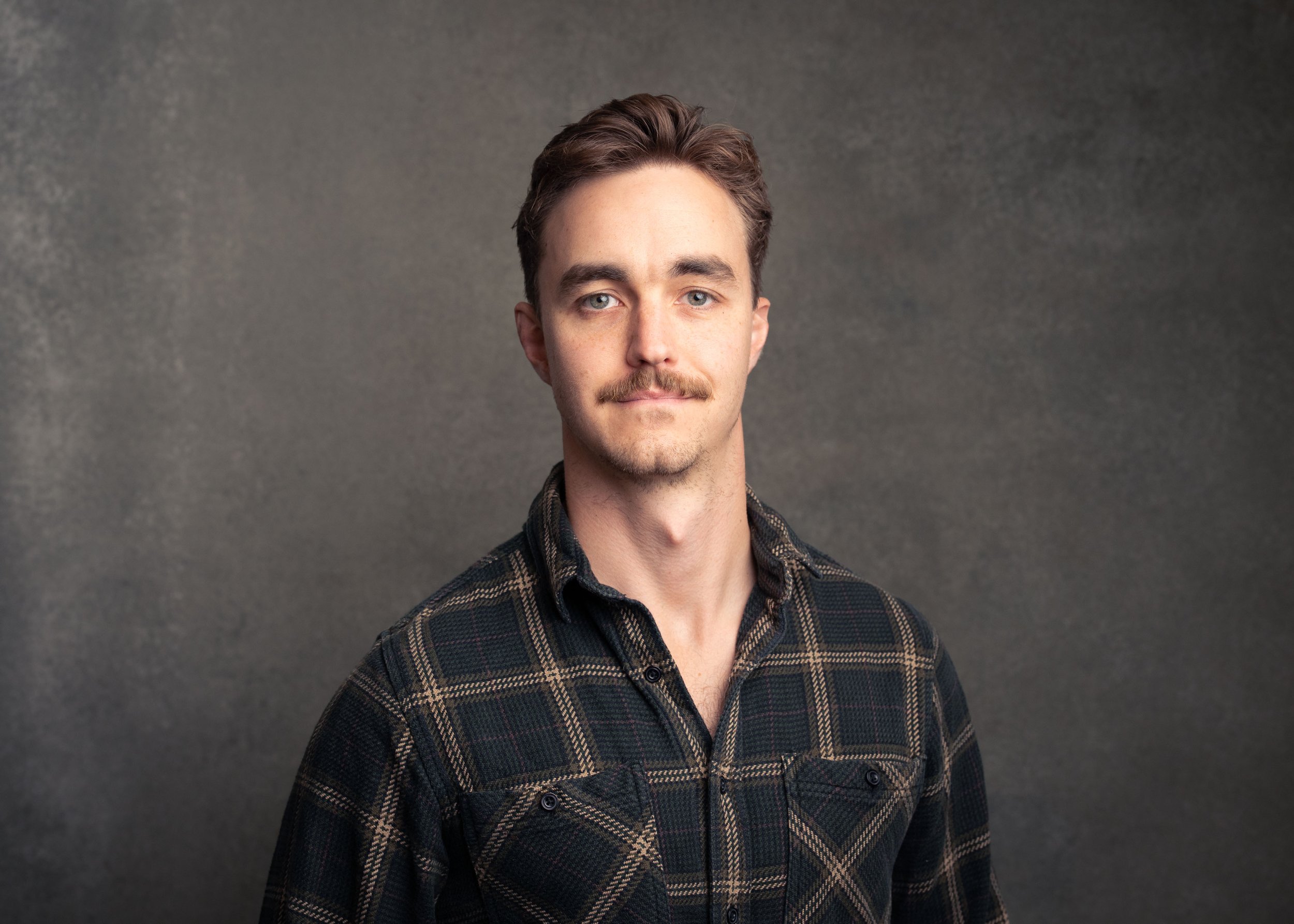 A man with brown hair, blue eyes, and a mustache wearing a black and brown plaid shirt against a plain gray background.