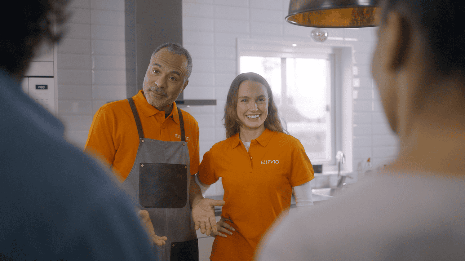 Two people in orange uniforms with 'ELLEVIO' logo talking to two people with blurred faces in a modern kitchen.