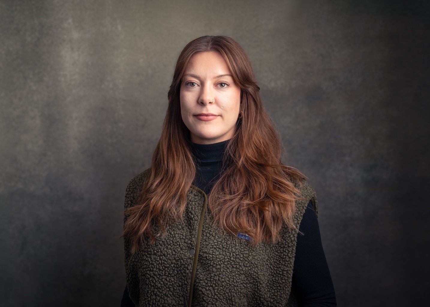 Portrait of a woman with long, wavy, reddish-brown hair wearing a black turtleneck and a green, fleece vest, standing against a dark, textured background.