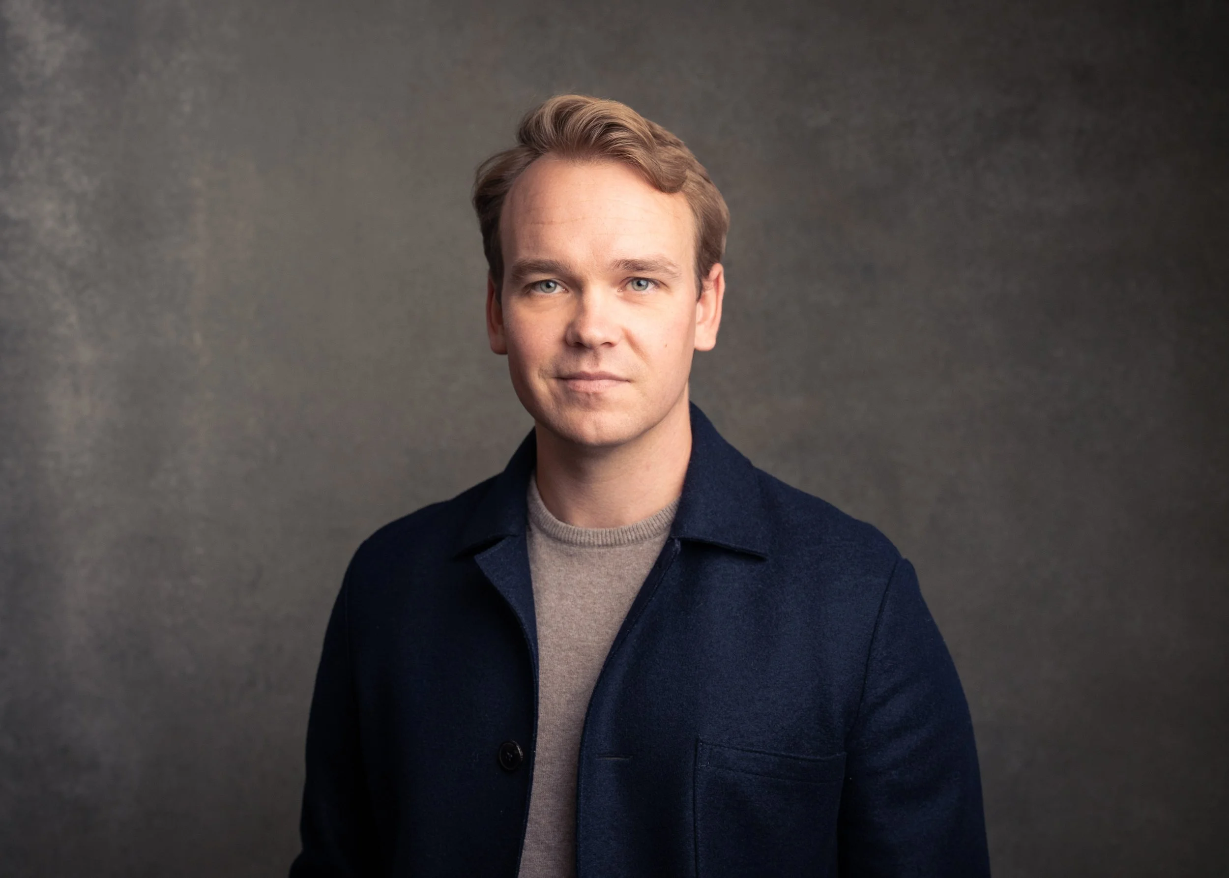 A man with light hair and blue eyes, wearing a gray t-shirt and a dark blazer, standing against a gray textured background.
