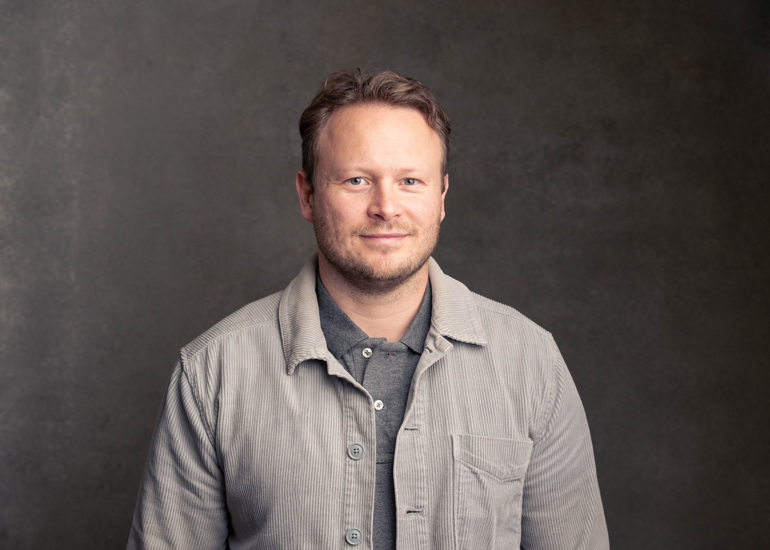 Portrait of a man with short brown hair, light beard, wearing a gray jacket and dark shirt, against a dark gray background.