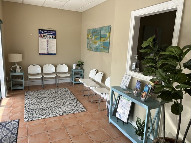 Waiting room with four white chairs, a small blue table, a patterned rug, a bulletin board, a large green houseplant, and a window with a hand sanitizer bottle on the window sill. A therapy room at The Speech Clinic of the Coastal Empire.