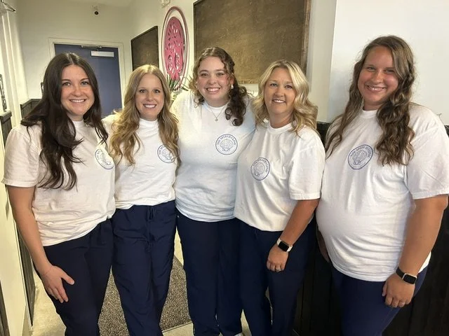 Five young women standing together indoors, smiling, wearing matching white t-shirts and navy blue pants. Therapists at The Speech Clinic of the Coastal Empire.