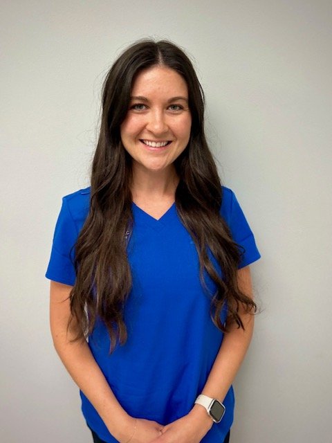 Young woman with long dark hair wearing blue medical scrubs, smiling, standing against a plain light gray wall.  A therapist with The Speech Clinic of the Coastal Empire in Savannah GA.