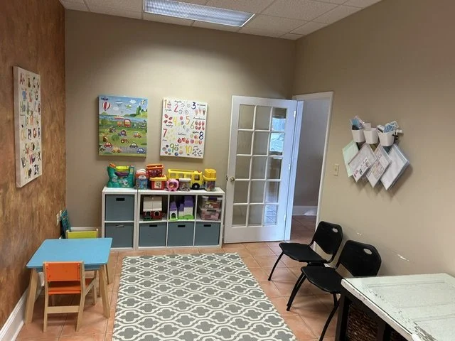 Playroom with small blue table and orange chair, storage unit with colorful toys, educational posters on the wall, black chairs, and a white door leading to another room. A therapy room at The Speech Clinic of the Coastal Empire.