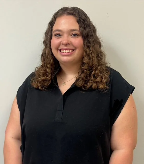 A young woman with curly brown hair, smiling and wearing a black top, standing against a plain light-colored wall. Speech Therapist in Pooler GA