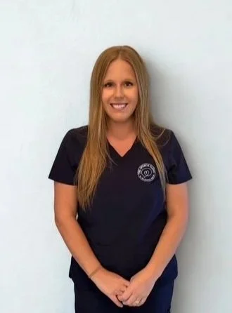 A woman with long blonde hair smiling, wearing navy scrubs, standing against a light blue wall. Adult speech therapy Pooler GA