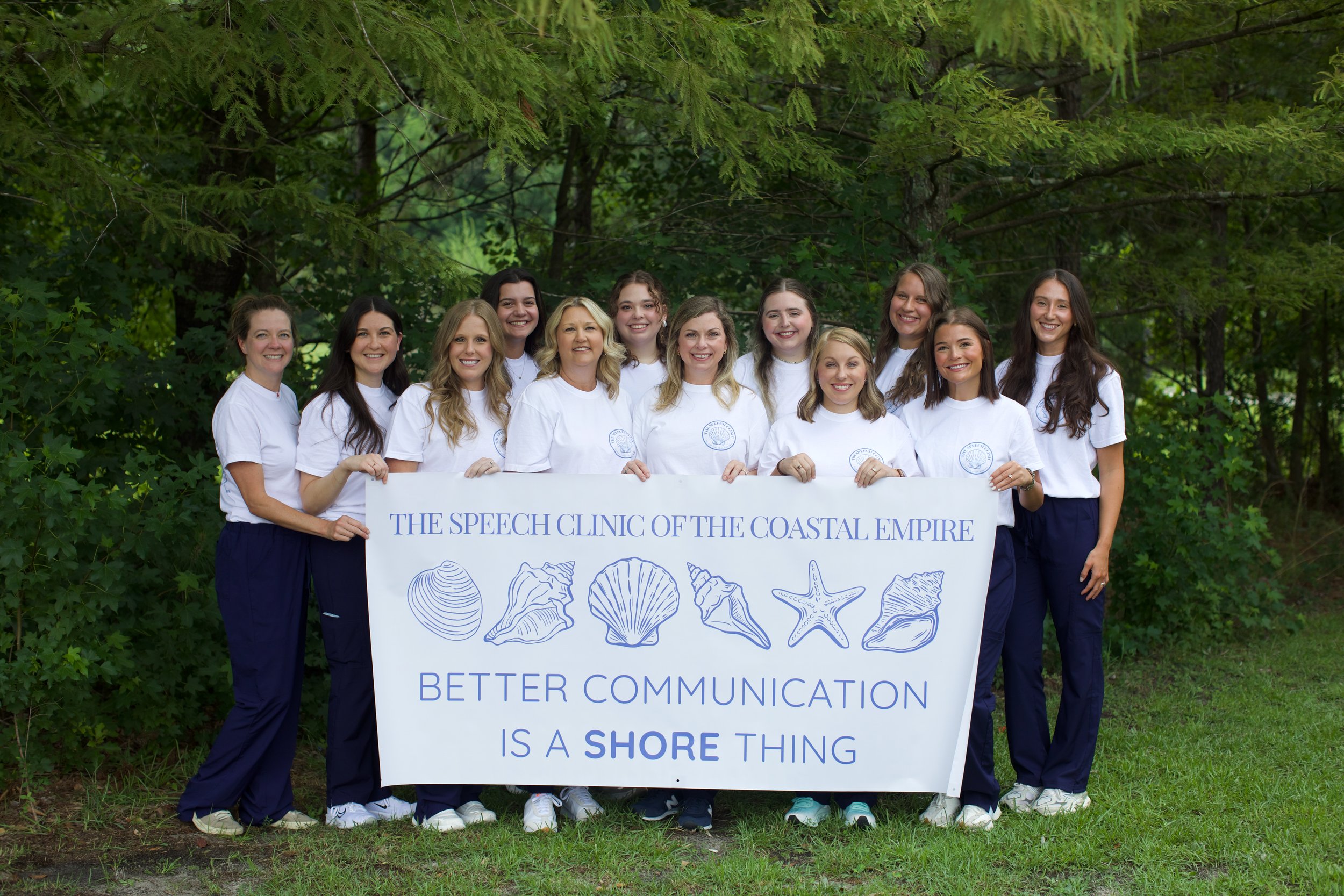 A group of eleven women standing outdoors under green trees, holding a large white banner with blue text and illustrations of seashells and starfish. The women are smiling and wearing white t-shirts and dark pants. The banner reads: 'The Speech Clinic of the Coastal Empire' and 'Better Communication is a Shore Thing'.
