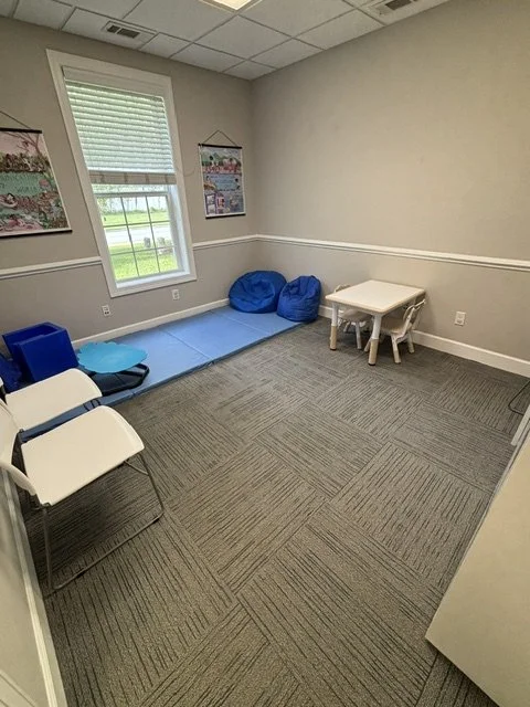 Empty room with a window, two chairs, a small table, two bean bags, and a padded area with a blue mat. A therapy room at The Speech Clinic of the Coastal Empire.